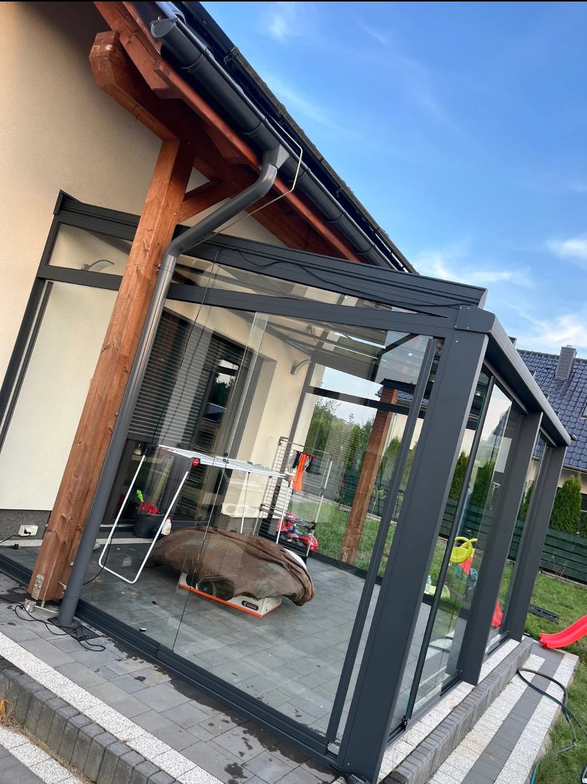 Szklana zabudowa tarasu z szarymi ramami, widok z zewnątrz. Wewnątrz suszarka na pranie i inne przedmioty. Kostka brukowa na tarasie. Błękitne niebo w tle.