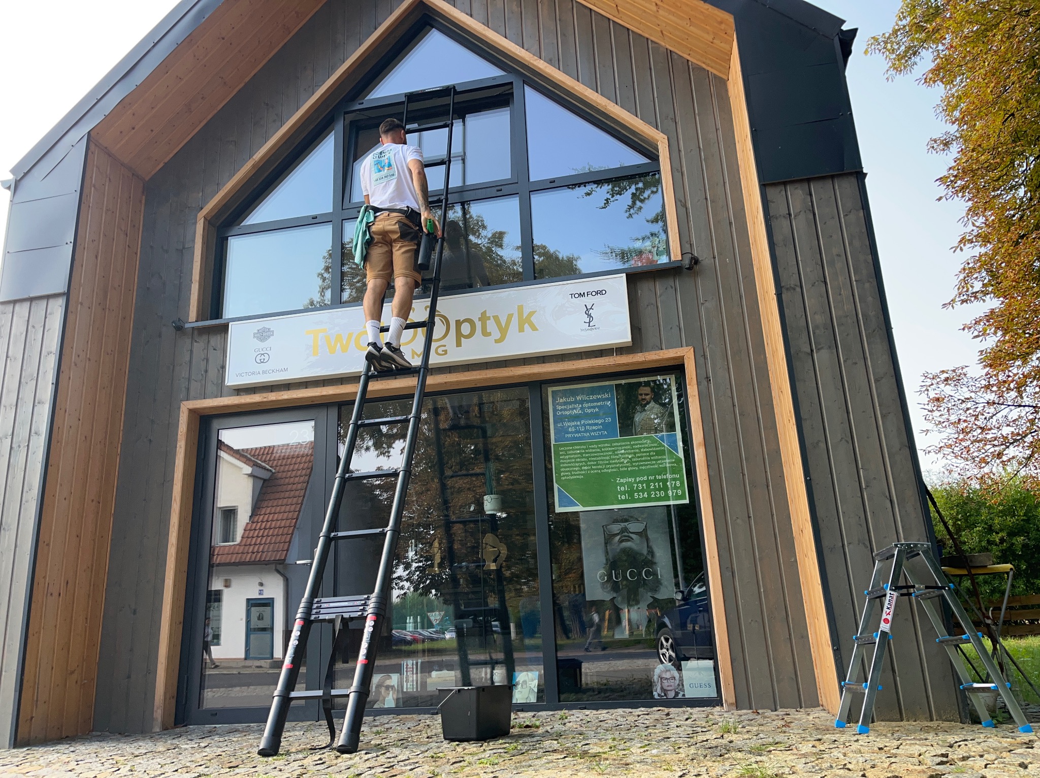 Mężczyzna na drabinie myje okna witryny salonu optycznego z logotypami luksusowych marek. Budynek o nowoczesnej architekturze z elementami drewna.