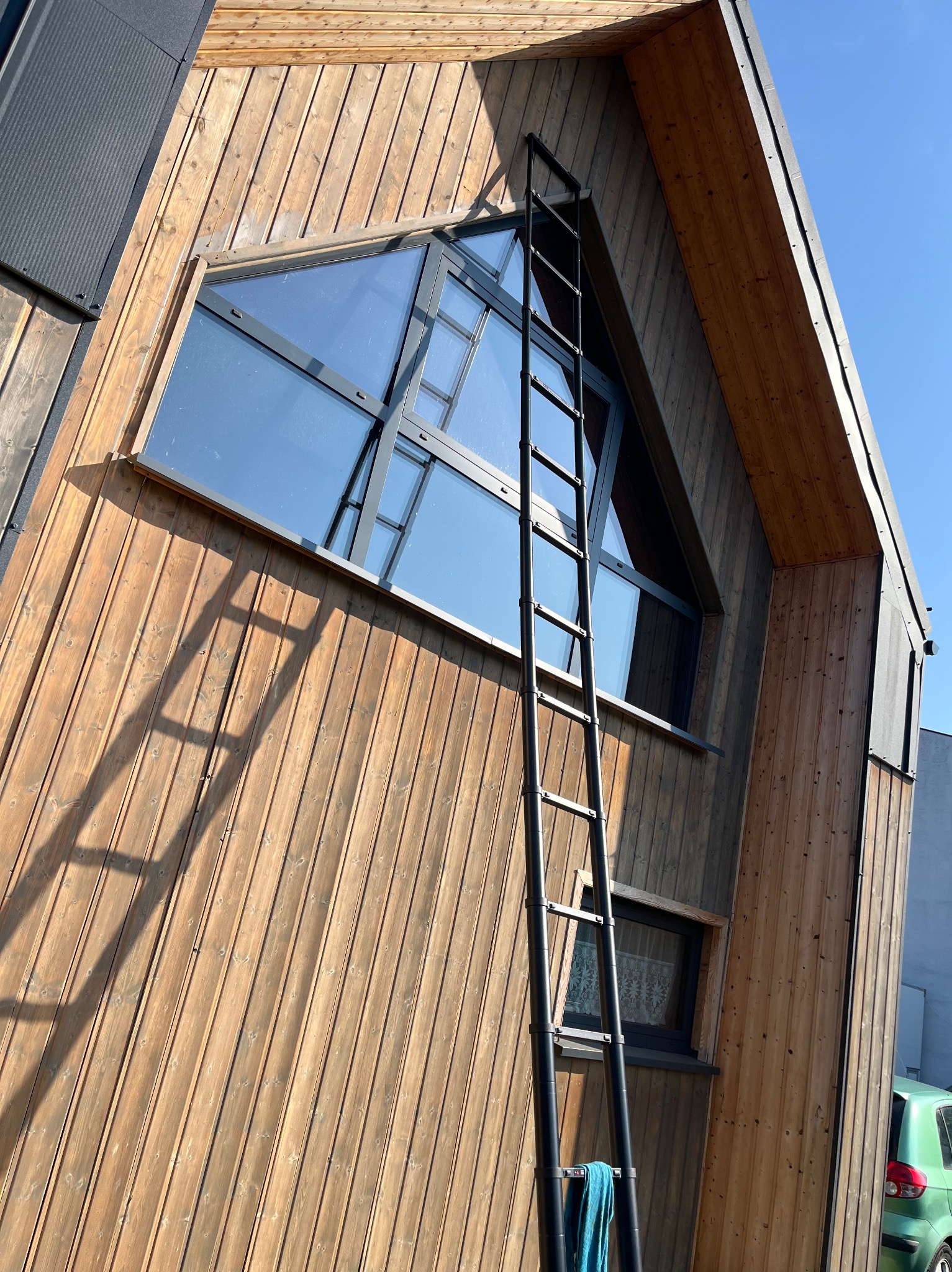 Elewacja domu z drewnianą okładziną i oknem w kształcie trójkąta. Przystawiona drabina teleskopowa z niebieską ścierką. Widoczne odbicie nieba w szybach.