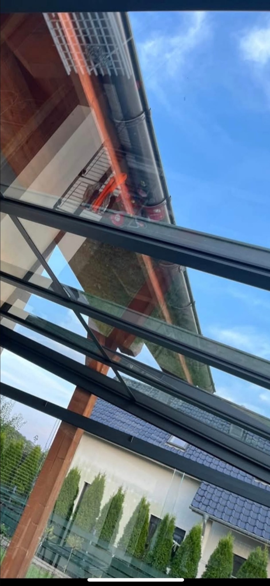 Szklany dach z widocznym zanieczyszczeniem i odbiciem nieba, wymagający czyszczenia. Widoczne rynny i otoczenie domu.