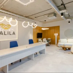 Nowoczesne wnętrze biura z długim białym stołem konferencyjnym, niebieskimi krzesłami i minimalistycznym oświetleniem. W tle sofa i drewniane panele ścienne.