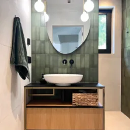Nowoczesna łazienka z umywalką nablatową, zielonymi płytkami na ścianie i drewnianą szafką. Minimalistyczne oświetlenie i lustro w owalnej ramie dodają elegancji.