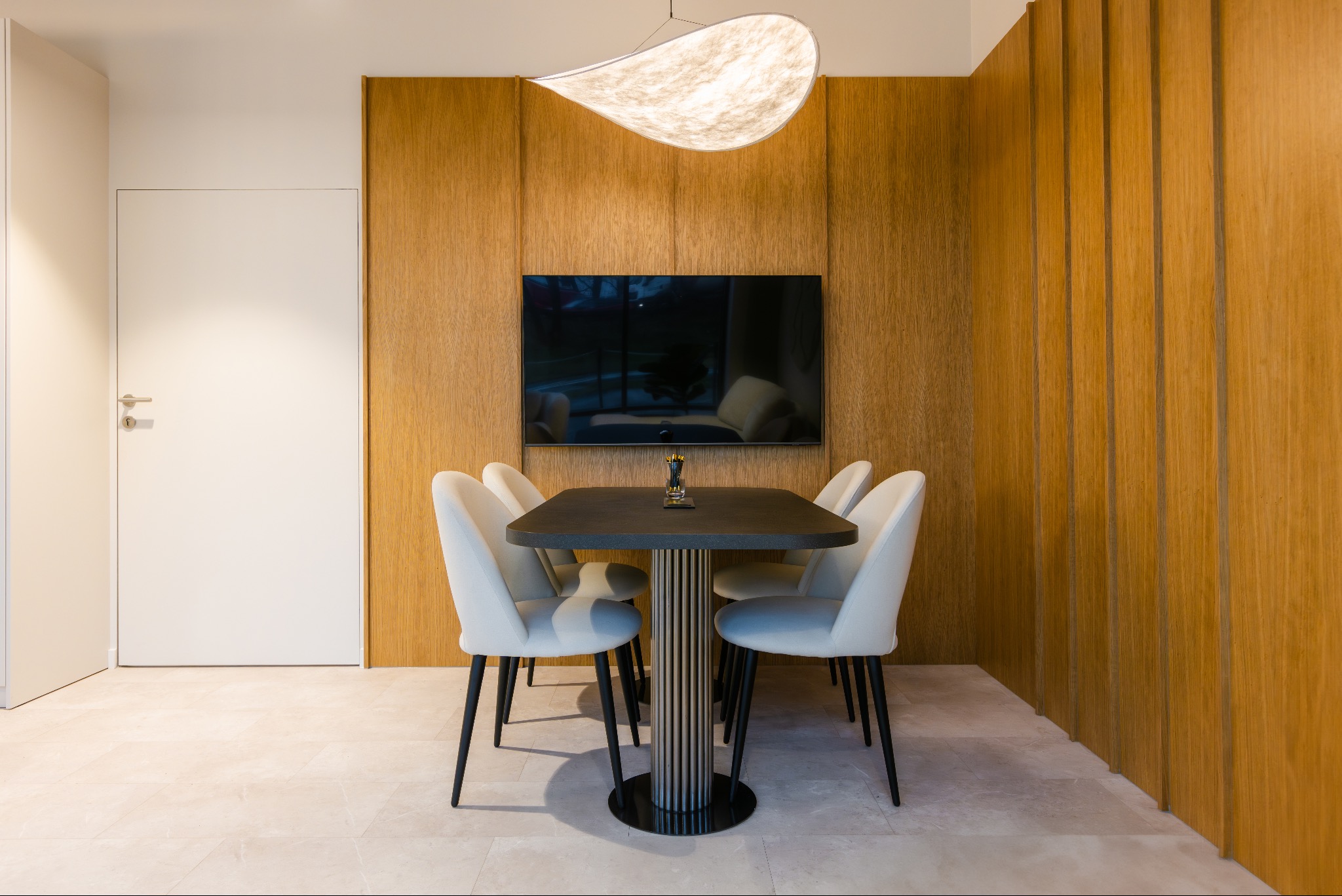 Jadalnia z drewnianą ścianą, stołem z ciemnym blatem, białymi krzesłami i designerską lampą. Telewizor na ścianie. Nowoczesna aranżacja wnętrza.