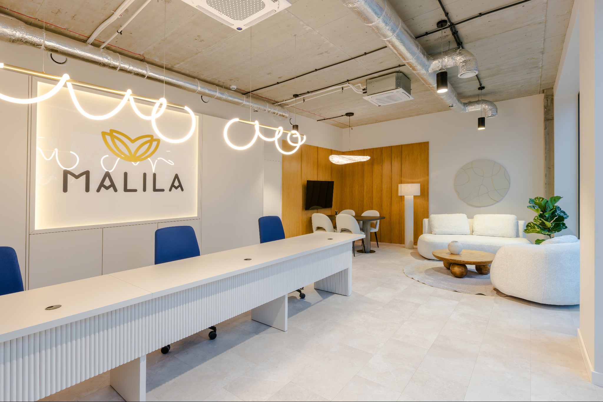 Nowoczesne wnętrze biura z długim białym stołem konferencyjnym, niebieskimi krzesłami i minimalistycznym oświetleniem. W tle sofa i drewniane panele ścienne.