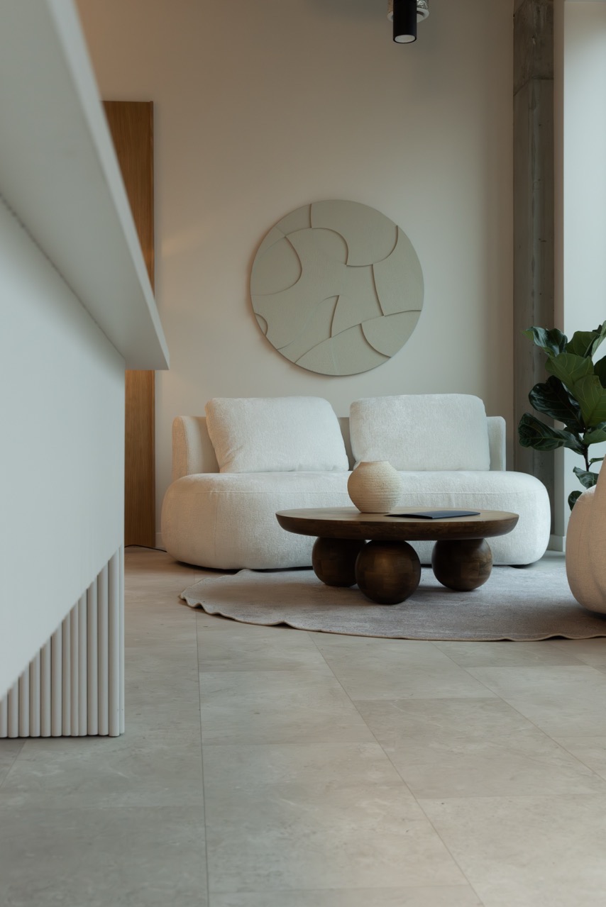 Minimalistyczny salon w neutralnych barwach: puszysta sofa, okrągły stolik kawowy z drewnianym blatem i ceramicznym wazonem, abstrakcyjna dekoracja ścienna.
