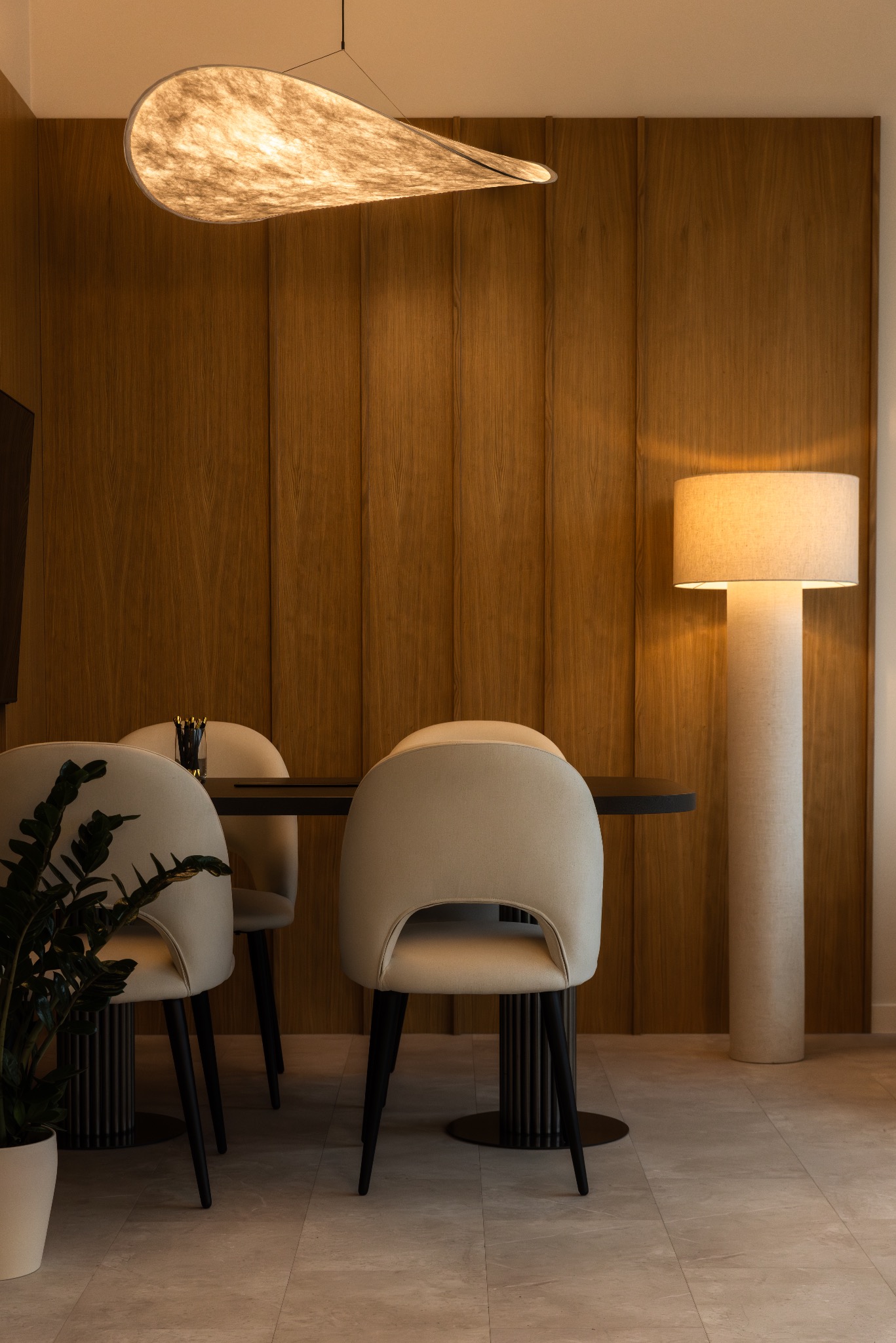 Minimalistyczna jadalnia: drewniana ściana, lampa w kształcie liścia, beżowe krzesła i czarny stół. Stonowana kolorystyka i nowoczesny design.