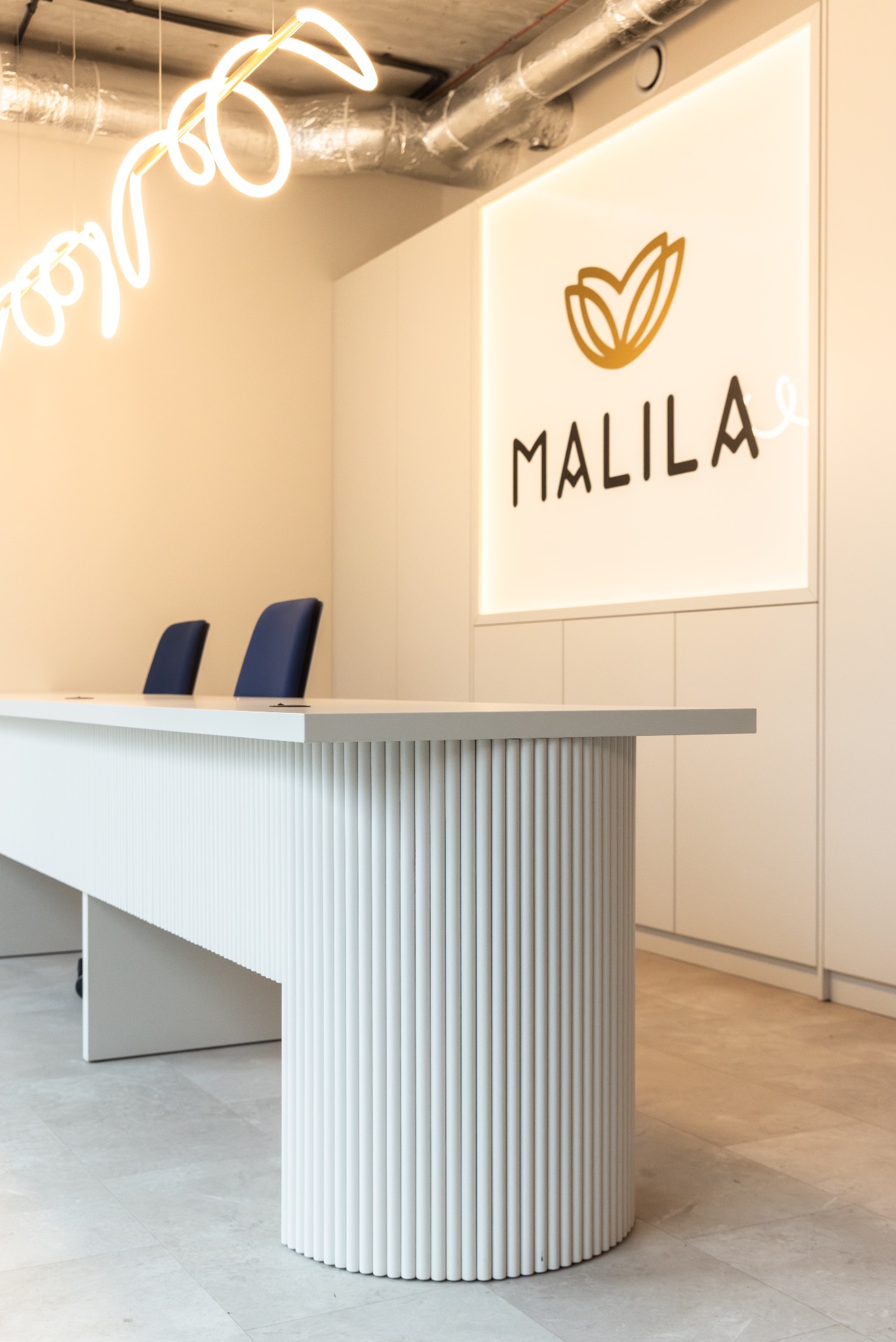 Minimalistyczne biuro z białą ladą recepcyjną o żłobkowanej fakturze, podświetlanym logo 'MALILA' i neonowym elementem dekoracyjnym pod sufitem.