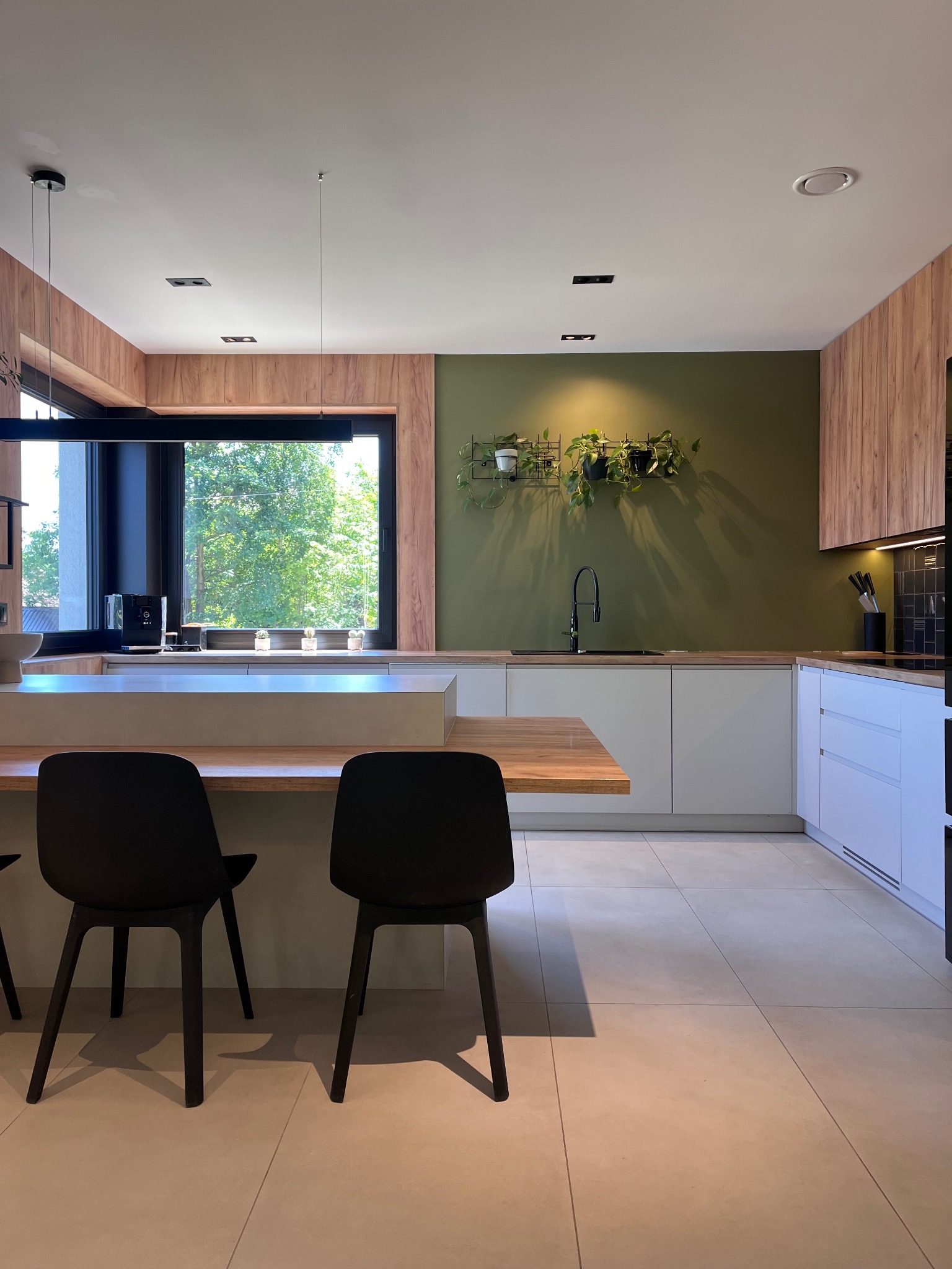 Nowoczesna kuchnia z wyspą i zieloną ścianą. Białe szafki, drewniane akcenty i czarne krzesła barowe. Widok na ogród przez duże okno.