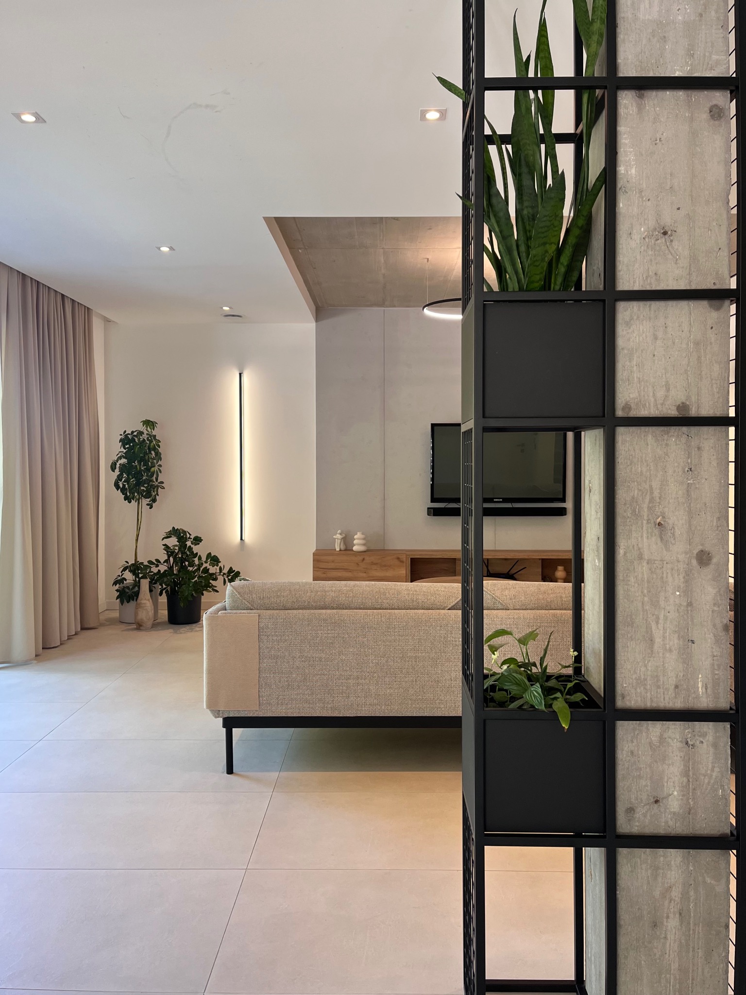 Nowoczesny salon z szarą sofą i regałem z betonu i metalu, w którym umieszczono rośliny. Minimalistyczne oświetlenie i jasne kolory tworzą spójną całość.