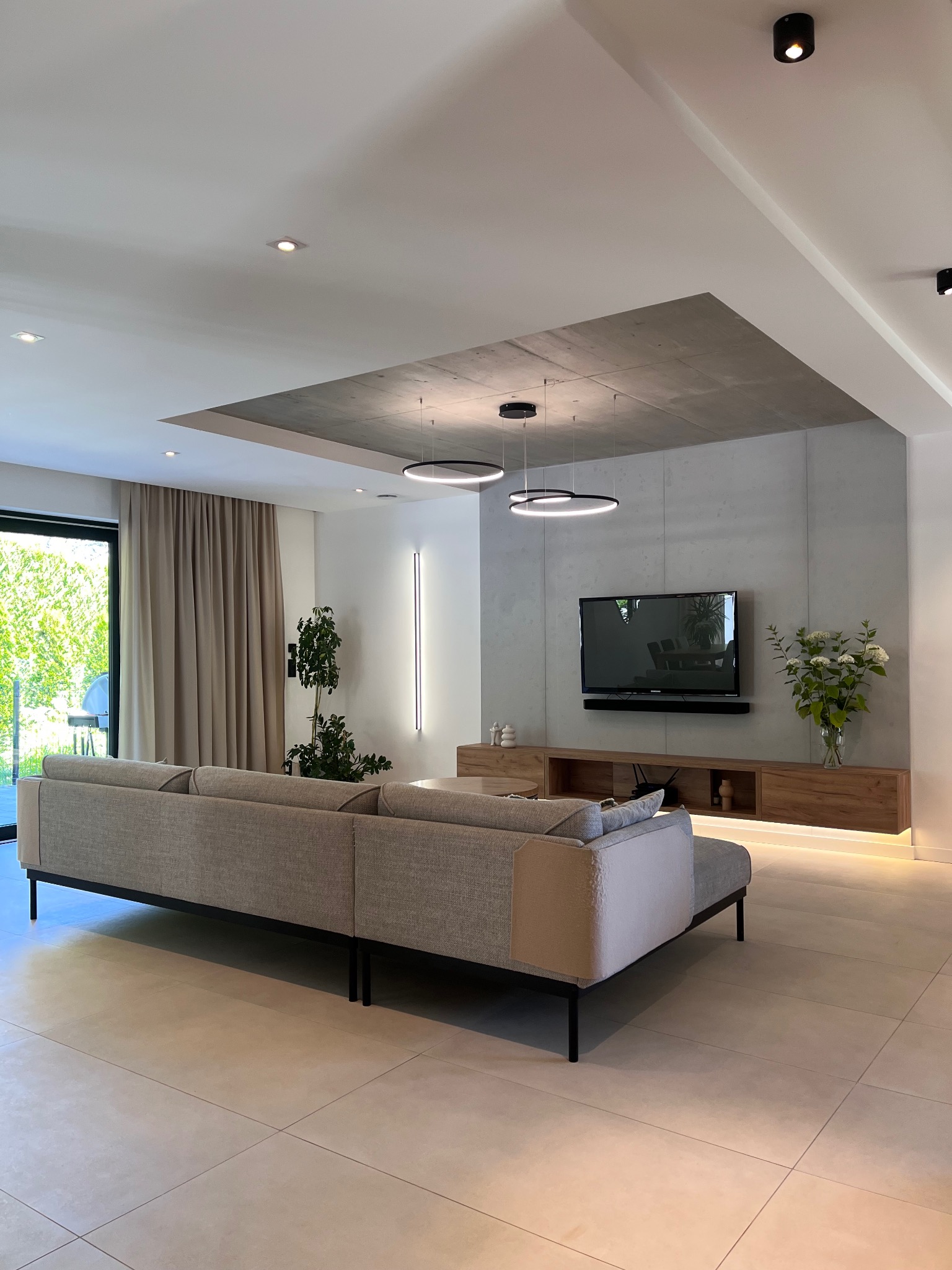 Nowoczesny salon z beżową sofą na tle betonowej ściany z telewizorem i drewnianą szafką RTV z podświetleniem LED. Okrągłe lampy wiszące nad sofą.