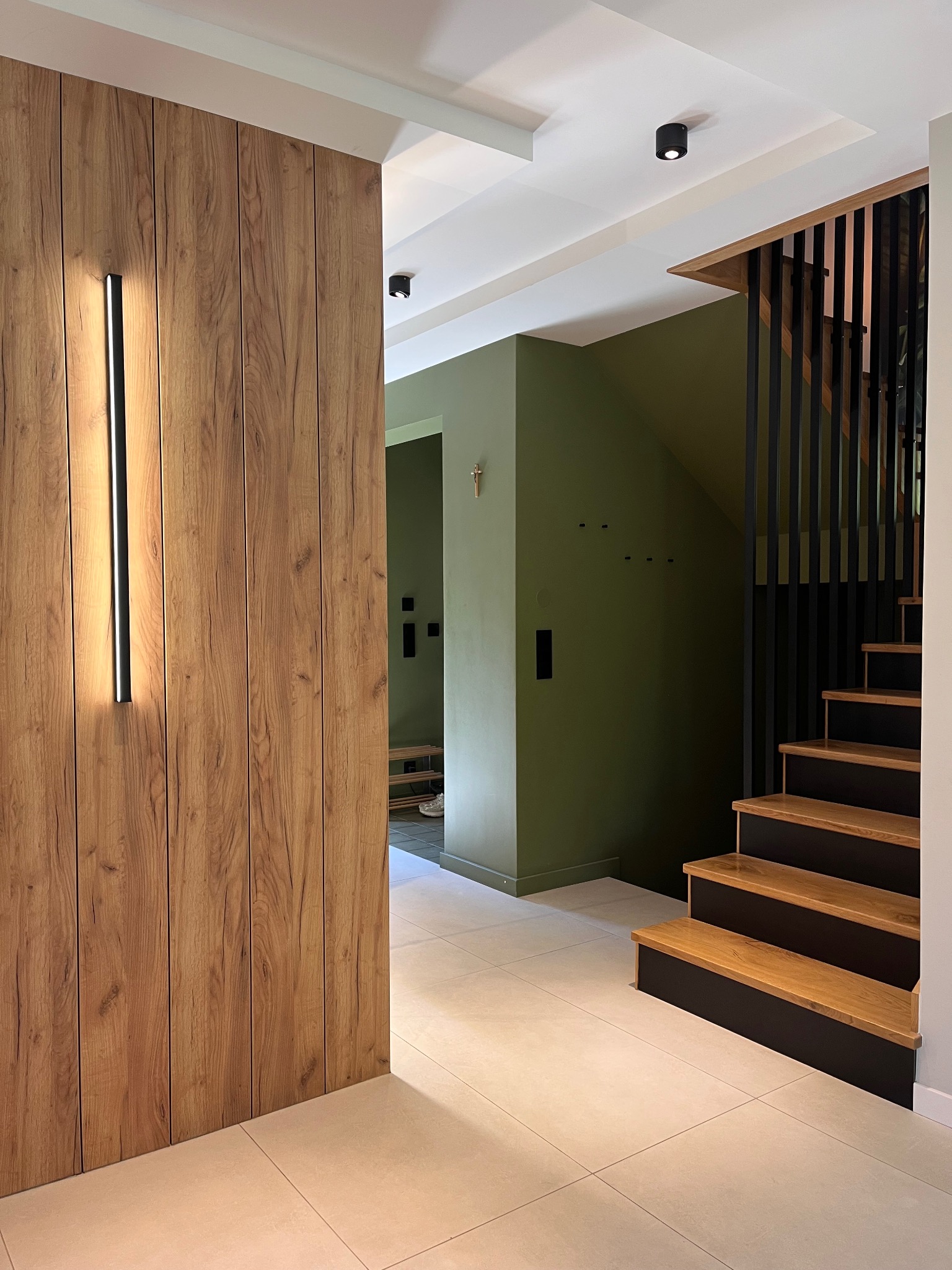 Nowoczesne wnętrze: drewniana ściana z oświetleniem, schody z czarną balustradą i oliwkowa ściana w tle. Minimalistyczny design z akcentami naturalnego drewna.
