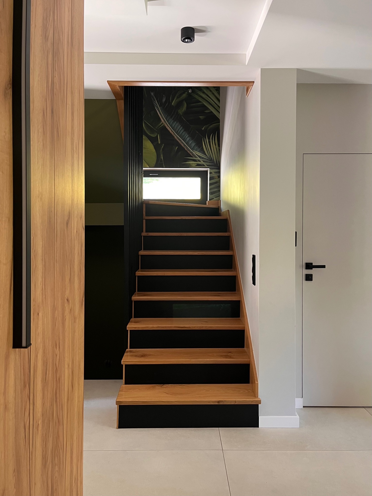 Nowoczesne schody z drewnianymi stopniami i czarnymi podstopnicami, oświetlone światłem z okna, z widoczną tapetą w tropikalne liście w tle.
