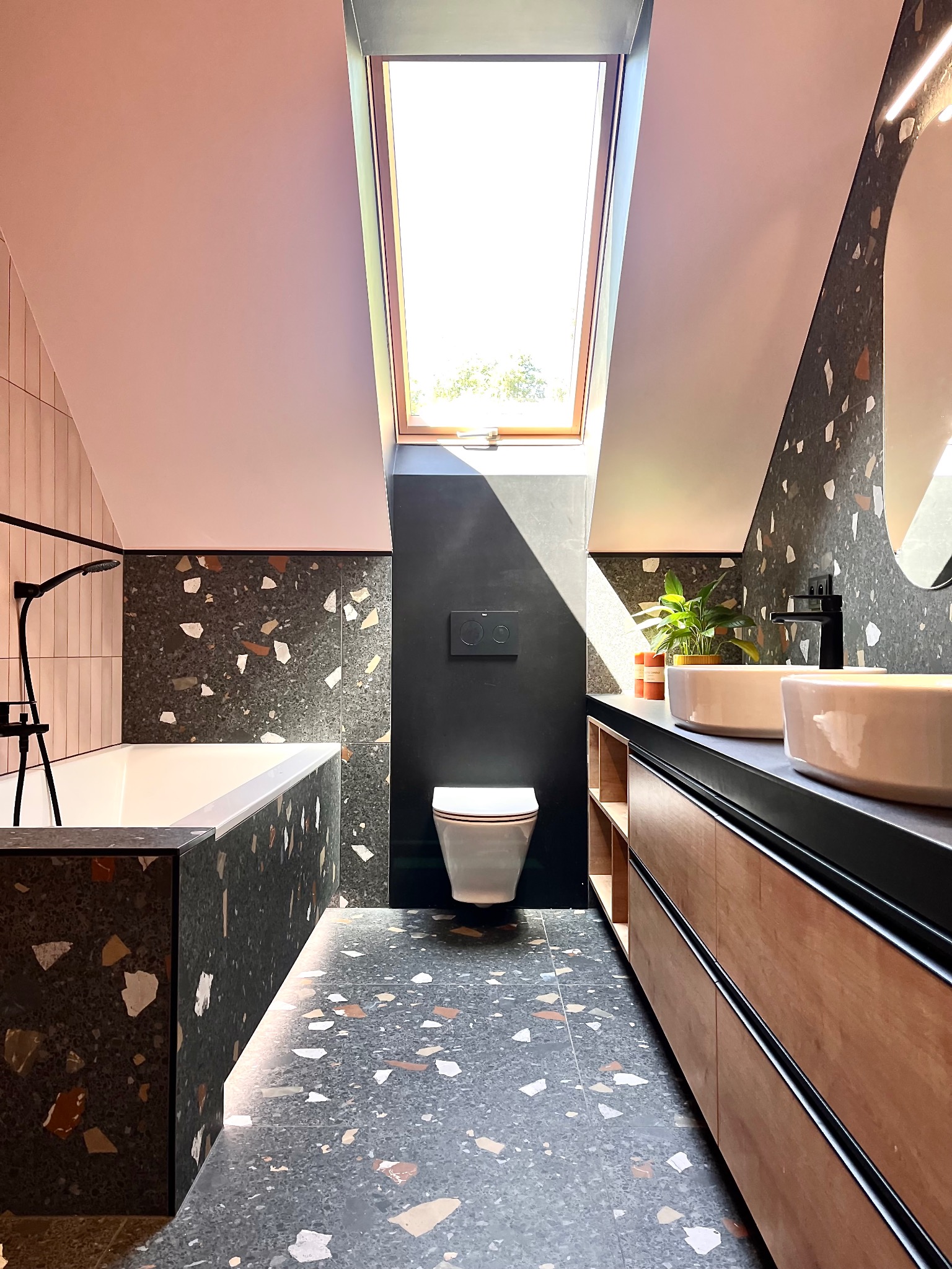 Nowoczesna łazienka na poddaszu z oknem dachowym, wykończona płytkami lastryko w odcieniach szarości i beżu, drewniane szafki pod umywalkami, minimalistyczna armatura.