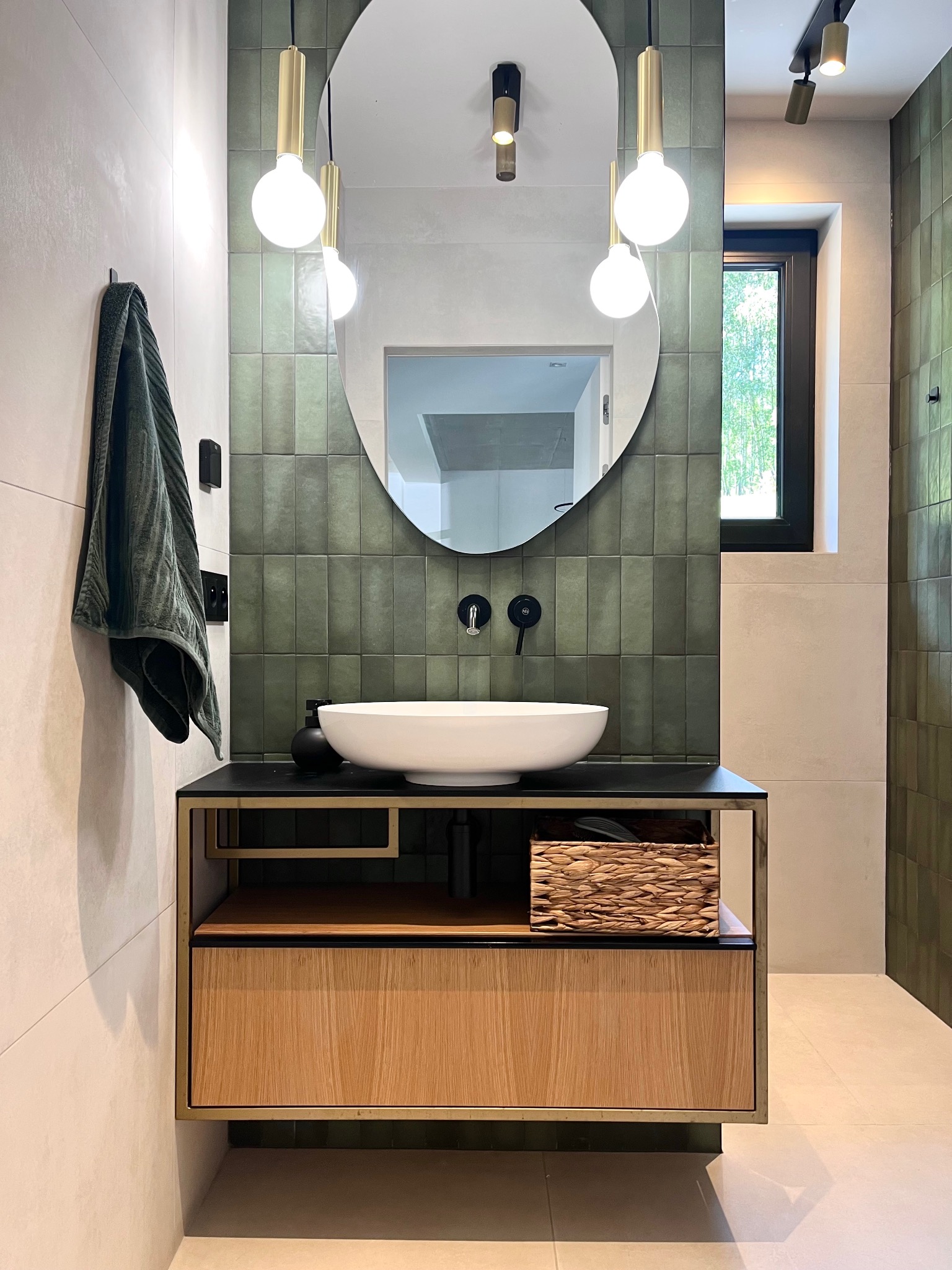 Nowoczesna łazienka z umywalką nablatową, zielonymi płytkami na ścianie i drewnianą szafką. Minimalistyczne oświetlenie i lustro w owalnej ramie dodają elegancji.