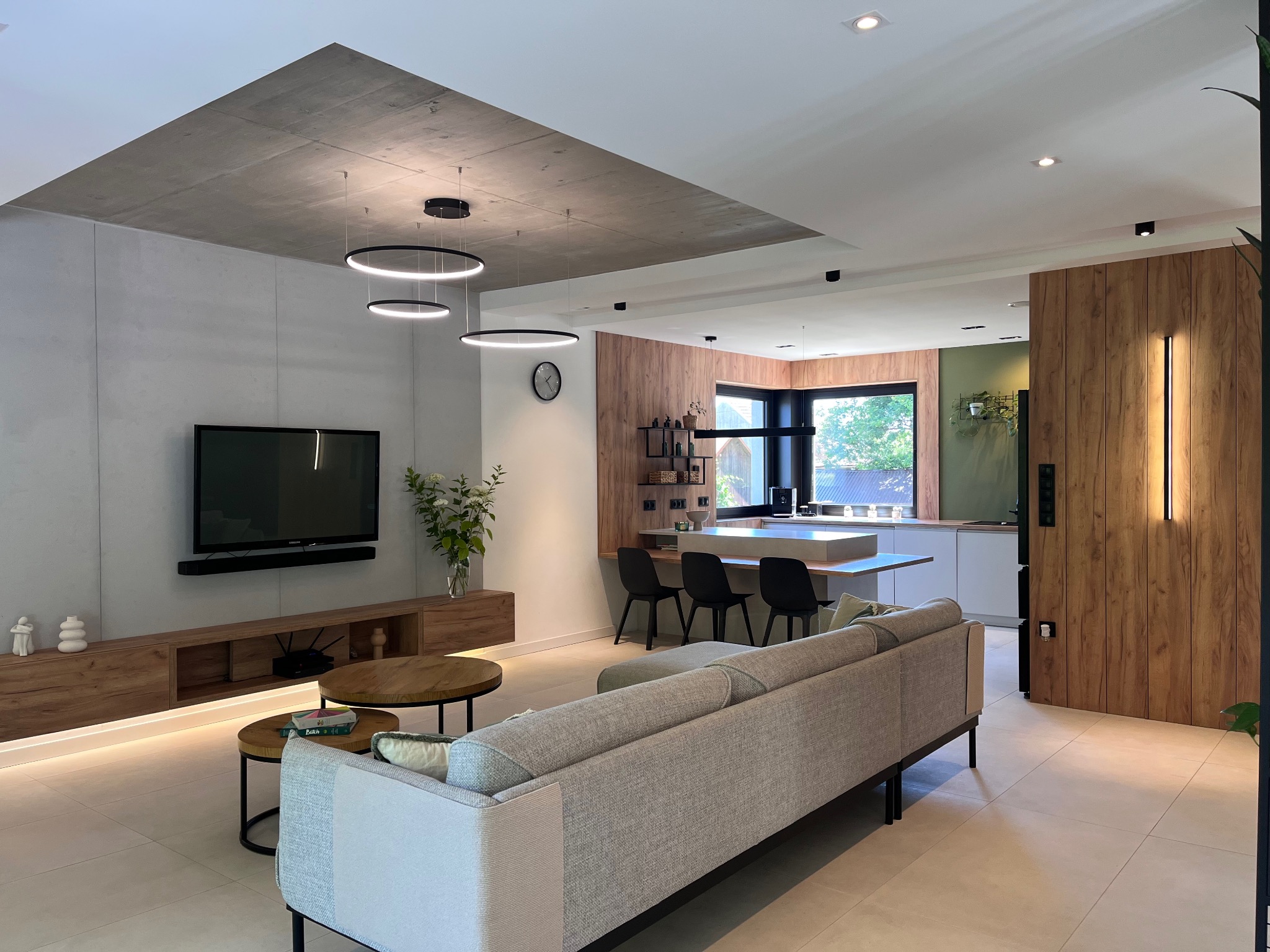 Nowoczesny salon z betonowym sufitem i drewnianymi panelami, oświetlony okrągłymi lampami. W tle widoczna kuchnia z oknem. Jasna podłoga i szara sofa.