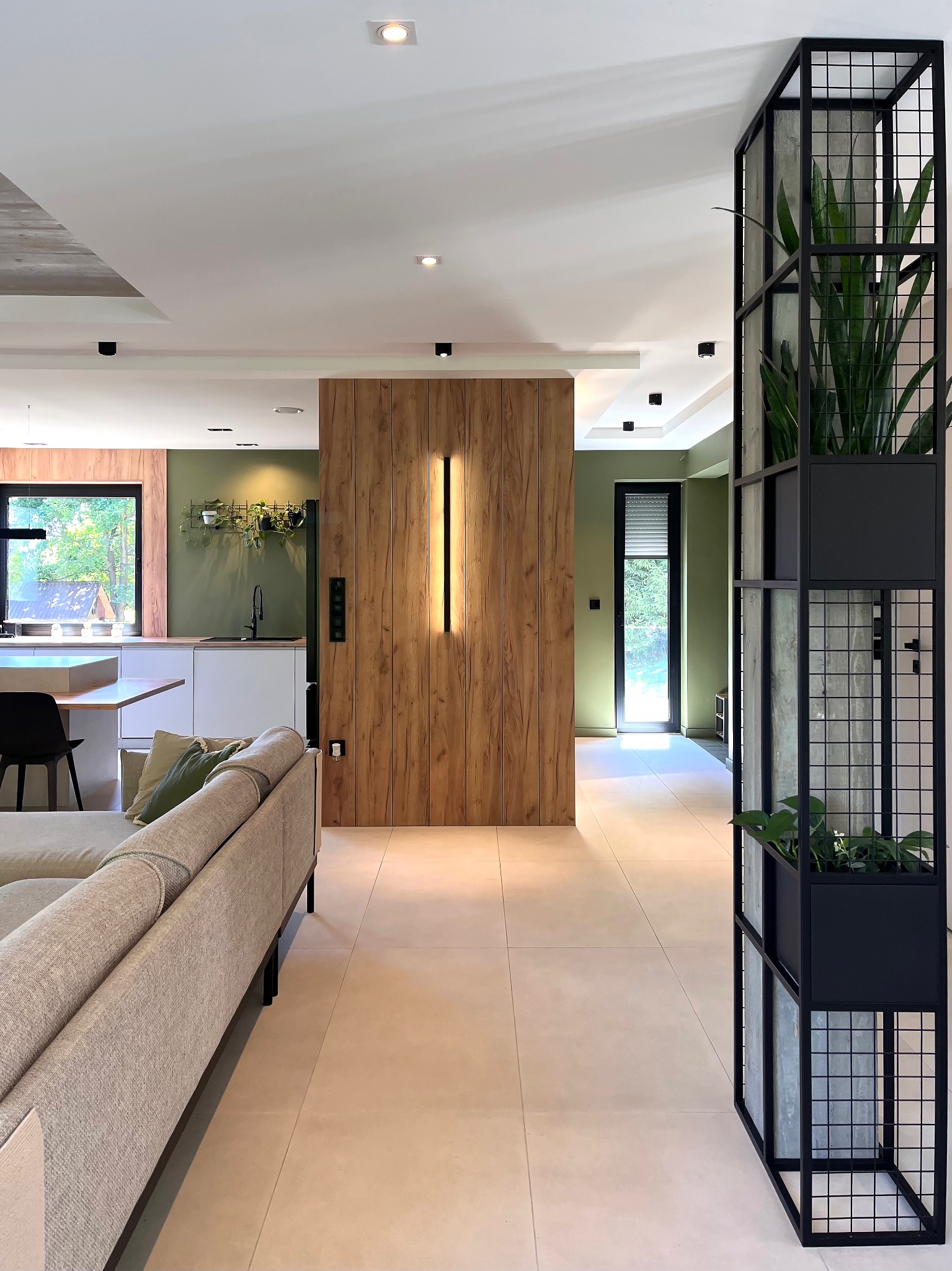 Nowoczesne wnętrze mieszkania z drewnianą ścianą i minimalistyczną metalową konstrukcją z roślinami, widok z perspektywy salonu w kierunku kuchni i okna.