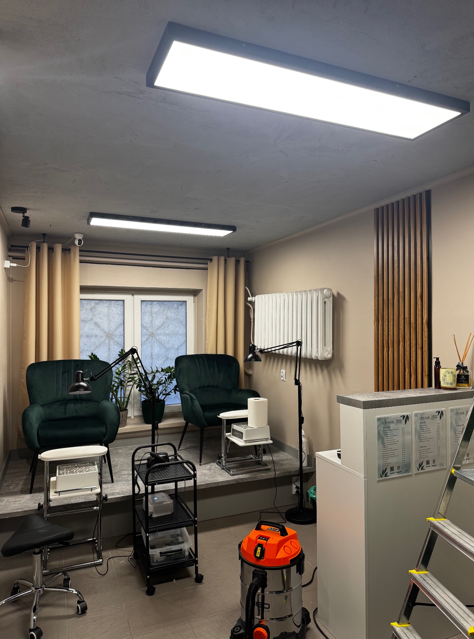 Wnętrze salonu kosmetycznego z oświetleniem sufitowym LED, dwoma fotelami, stanowiskami pracy i elementami dekoracyjnymi z drewna. W tle okno z zasłonami.