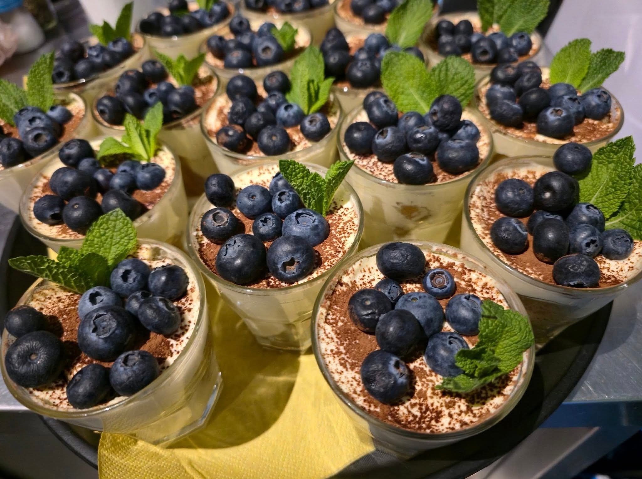Deser tiramisu w szklankach, posypany kakao i udekorowany świeżymi borówkami oraz listkiem mięty, serwowany na tacy. Elegancki catering.