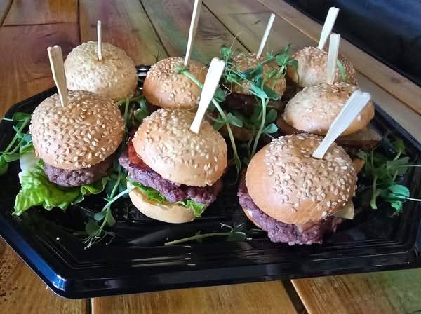 Mini burgery z sezamem na czarnej tacy, dekorowane zieleniną i patyczkami. Idealne na catering imprezowy, z bliska, na drewnianym tle.