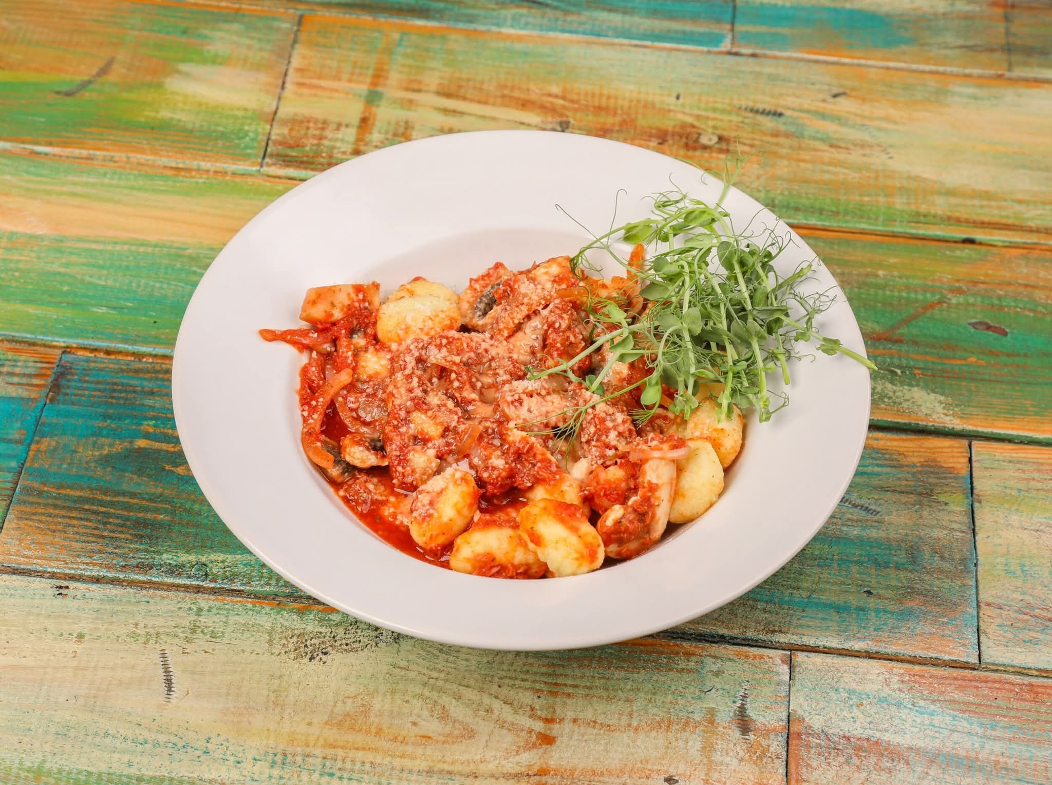 Gnocchi w sosie pomidorowym, posypane serem i udekorowane zielonymi pędami na białym talerzu, na tle rustykalnego, kolorowego stołu.