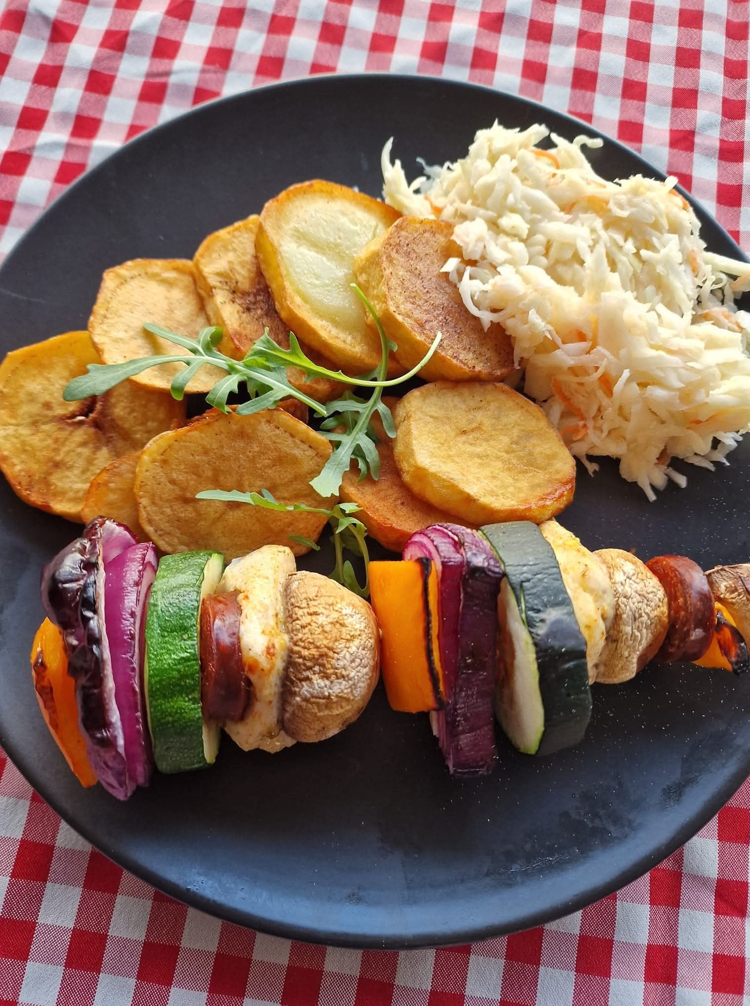 Szpadka z warzywami i kiełbasą, frytki z ziemniaków i surówka z kapusty na czarnym talerzu z rucolą, na czerwonej kraciastej ściereczce.