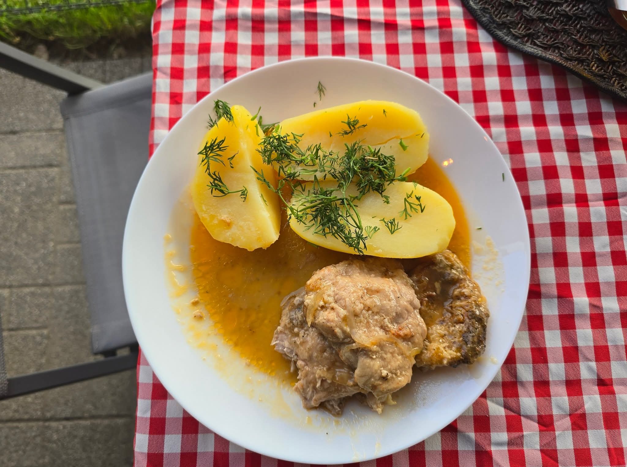 Porcja obiadowa na talerzu: ziemniaki z koperkiem i mięso w sosie, na czerwono-białym obrusie w kratę, obok krzesło ogrodowe. Domowy obiad.