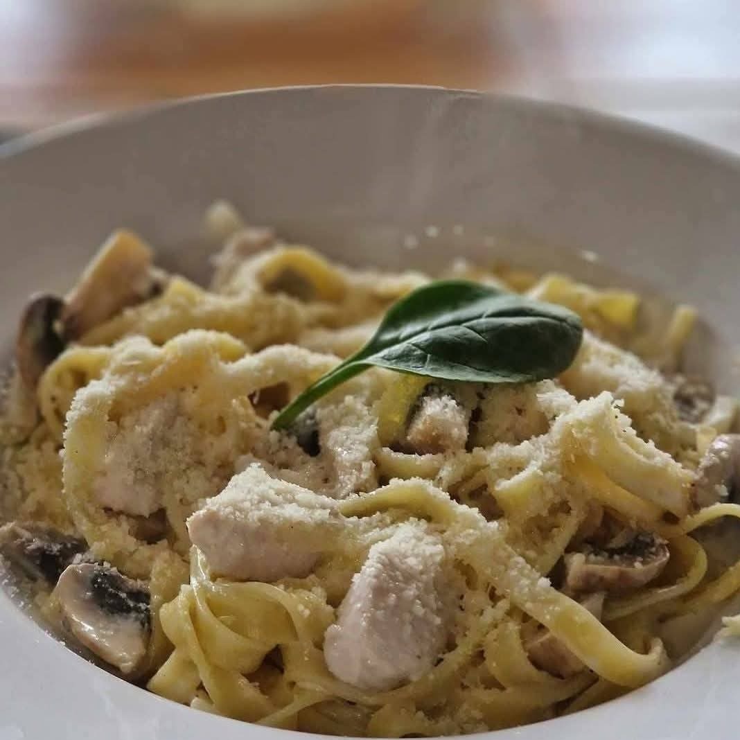 Makaron tagliatelle z kurczakiem, pieczarkami i parmezanem, udekorowany listkiem szpinaku, serwowany w białym, głębokim talerzu.