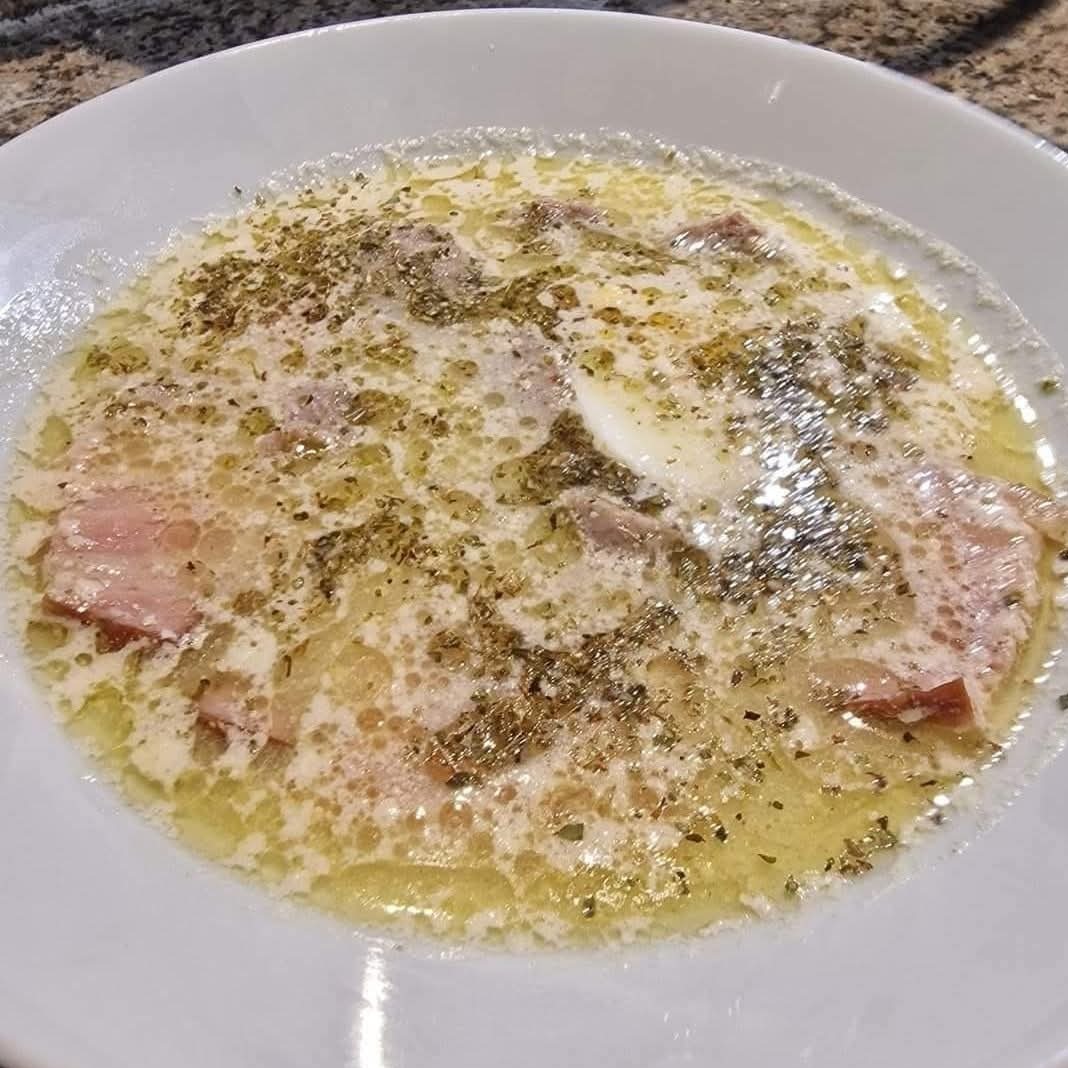 Żurek w białym talerzu z boczkiem, jajkiem i ziołami, z widocznymi pęcherzykami tłuszczu, na blacie z szarego, nakrapianego kamienia.