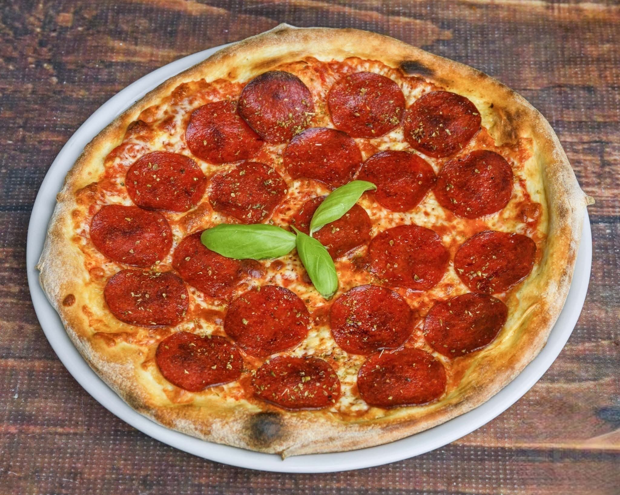 Apetyczna pizza pepperoni na białym talerzu, z bazylią w centrum, na tle drewnianego stołu. Widok z góry, idealna propozycja na catering.