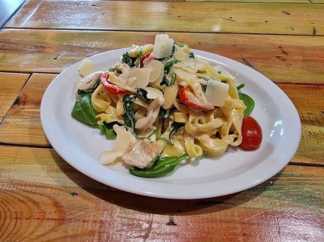 Tagliatelle z kurczakiem, szpinakiem, pomidorami i parmezanem na białym talerzu, na tle drewnianego stołu. Danie gotowe do podania.