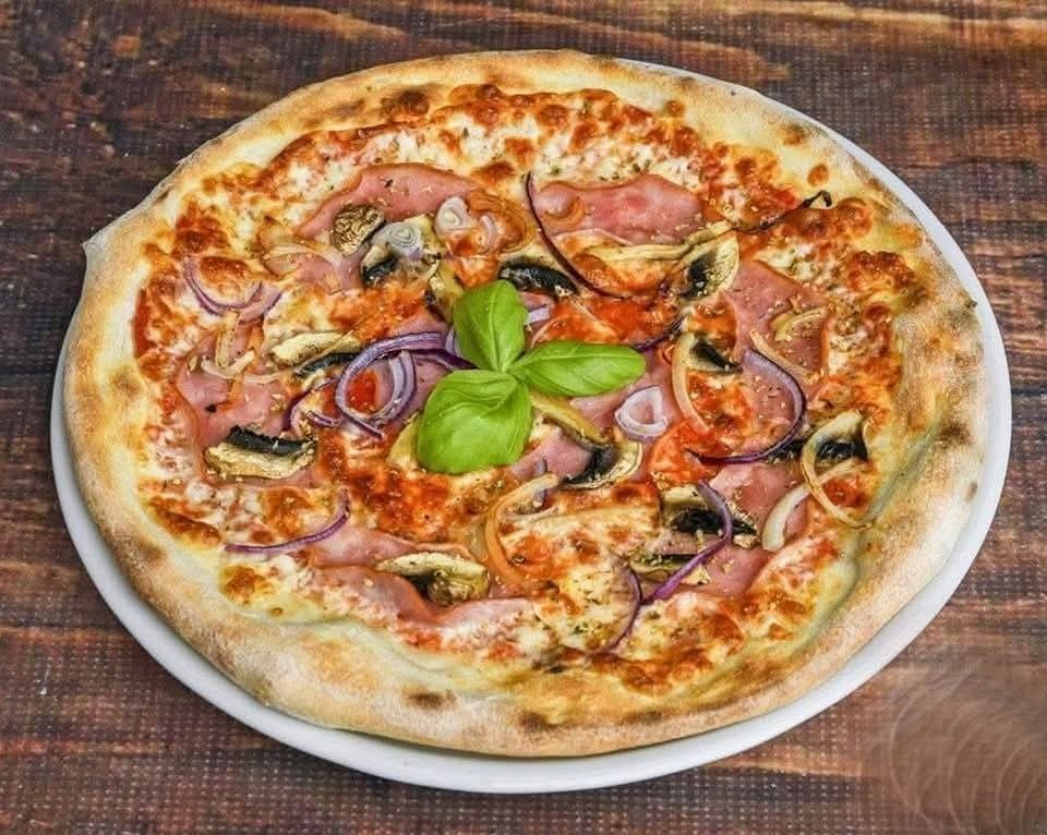 Apetyczna pizza z szynką, pieczarkami i czerwoną cebulą na cienkim cieście, udekorowana świeżą bazylią, serwowana na białym talerzu na drewnianym stole.