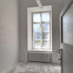 Pokój po malowaniu na biało, zabezpieczone okno folią, grzejnik i podłoga przykryte folią malarską. Widoczne świeżo pomalowane ściany i sufit.