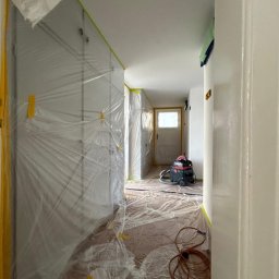 Maciej Antończyk FinishLab - Korytarz zabezpieczony folią malarską i taśmą przed malowaniem. Widoczny odkurzacz przemysłowy i przewód. Przygotowania do prac wykończeniowych.