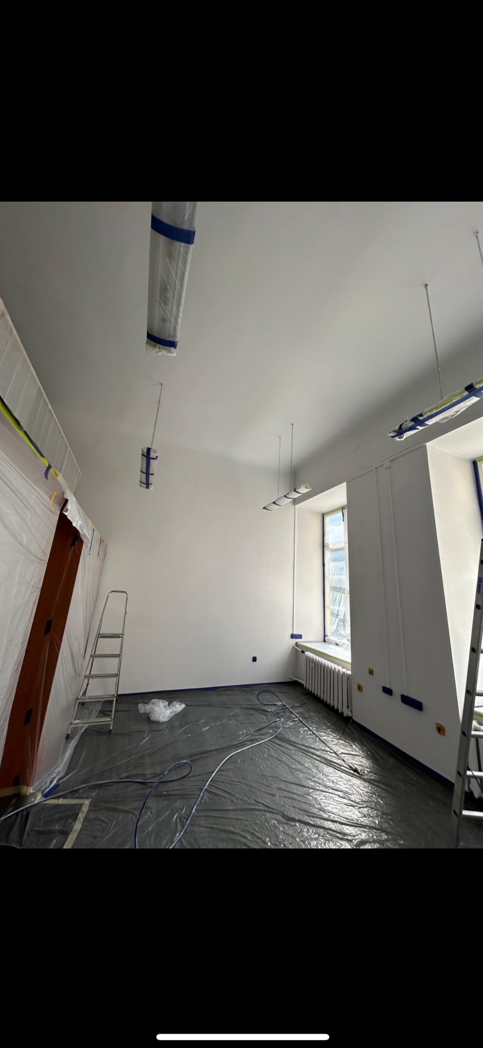 Pokój po malowaniu na biało, zabezpieczony folią i taśmą malarską, z widoczną drabiną i grzejnikiem pod oknem. Przygotowanie do prac wykończeniowych.