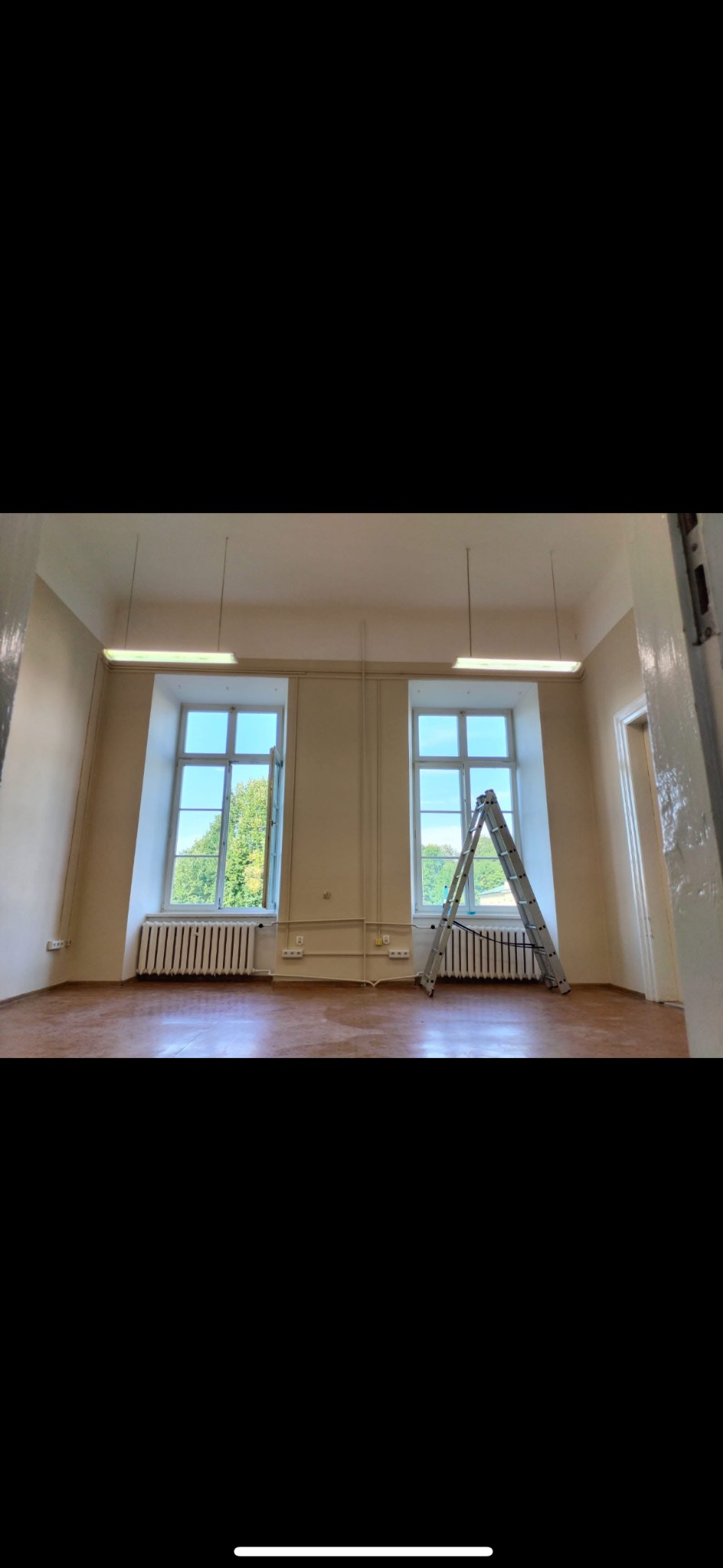 Wnętrze pokoju z dwoma oknami i drabiną opartą o grzejnik. Ściany pomalowane na jasny kolor, drewniana podłoga, lampy sufitowe. Przygotowanie do prac remontowych.