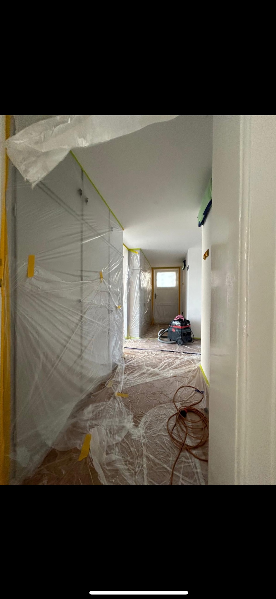 Korytarz zabezpieczony folią malarską i taśmą przed malowaniem. Widoczny odkurzacz przemysłowy i przewód. Przygotowania do prac wykończeniowych.