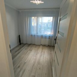 K.S bud - Jasne panele podłogowe w pustym pokoju z oknem zasłoniętym firaną. Widok przez otwarte drzwi z białymi panelami dekoracyjnymi. Na suficie nowoczesna lampa.