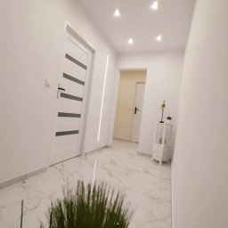 K.S bud - Biały korytarz z marmurową podłogą, oświetleniem punktowym i dekoracyjną rośliną. Widoczne drzwi z czarnymi akcentami i minimalistyczna komoda.