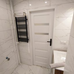 K.S bud - Elegancka łazienka z białymi marmurkowymi płytkami, czarnym grzejnikiem drabinkowym i białą stolarką drzwiową z czarną klamką. Minimalistyczny design.