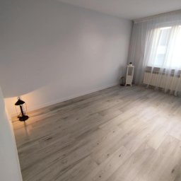 K.S bud - Jasne panele podłogowe w pustym pokoju z białymi ścianami i oknem z firanką. W rogu stoi lampka, a przy oknie mała szafka. Minimalistyczne wykończenie wnętrza.