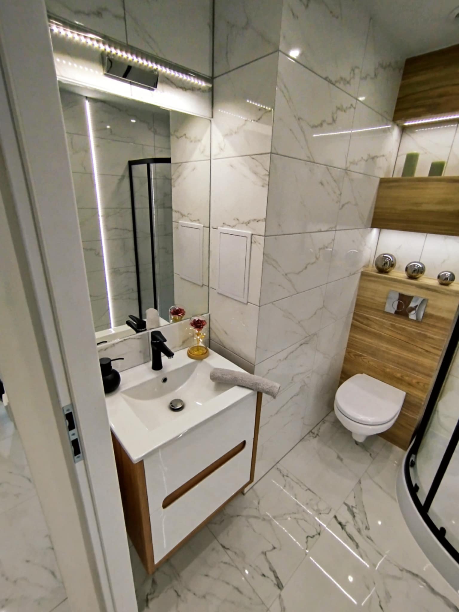 Nowoczesna łazienka z marmurowymi płytkami, podświetlanym lustrem, drewnianymi akcentami i białą armaturą. Widok na umywalkę, toaletę i fragment kabiny prysznicowej.