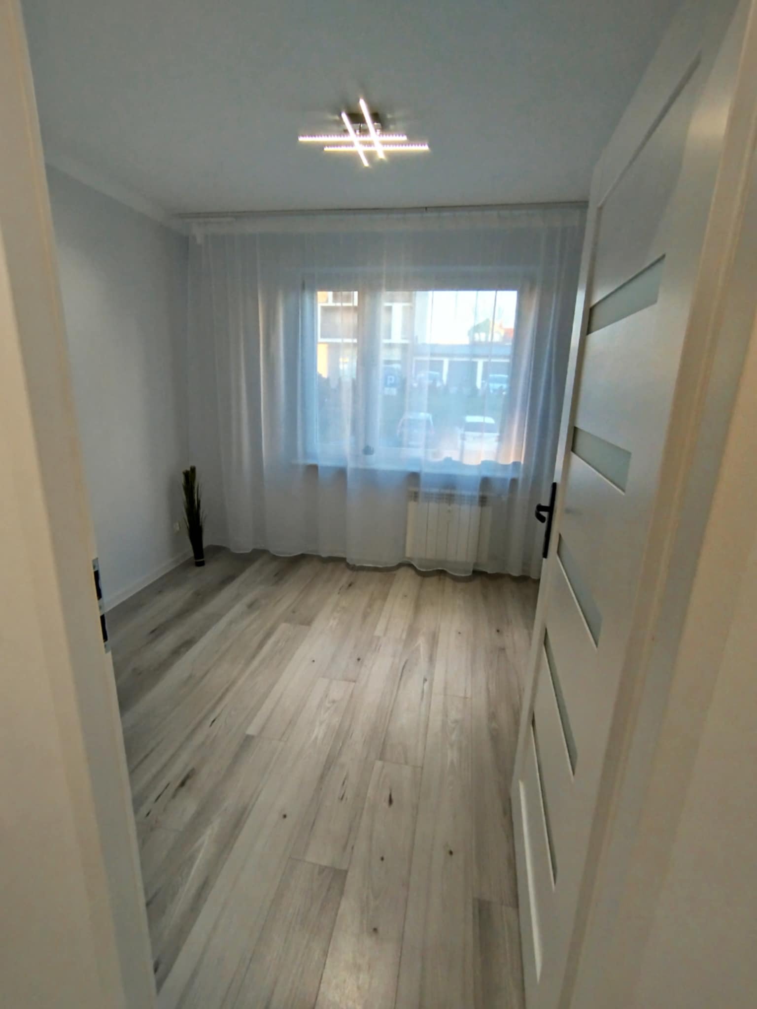 Jasne panele podłogowe w pustym pokoju z oknem zasłoniętym firaną. Widok przez otwarte drzwi z białymi panelami dekoracyjnymi. Na suficie nowoczesna lampa.