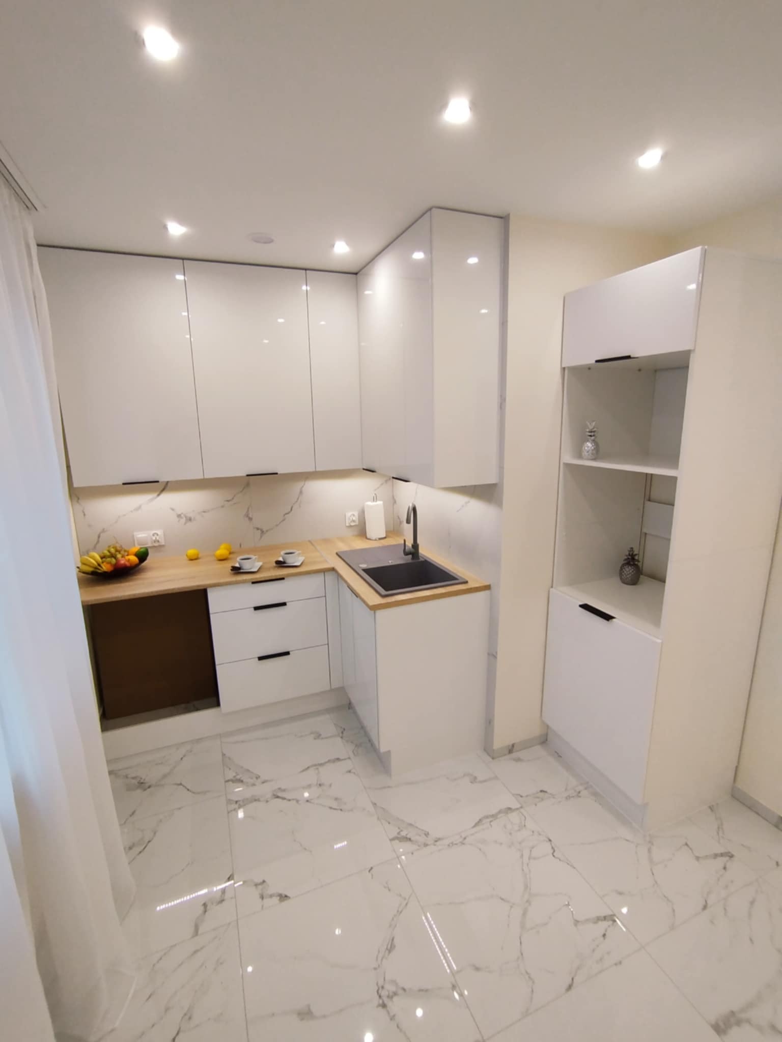 Biała, nowoczesna kuchnia z marmurową podłogą i drewnianymi blatami. Widoczne owoce, filiżanki, szafki z czarnymi uchwytami i dekoracyjne ananasy na półkach.