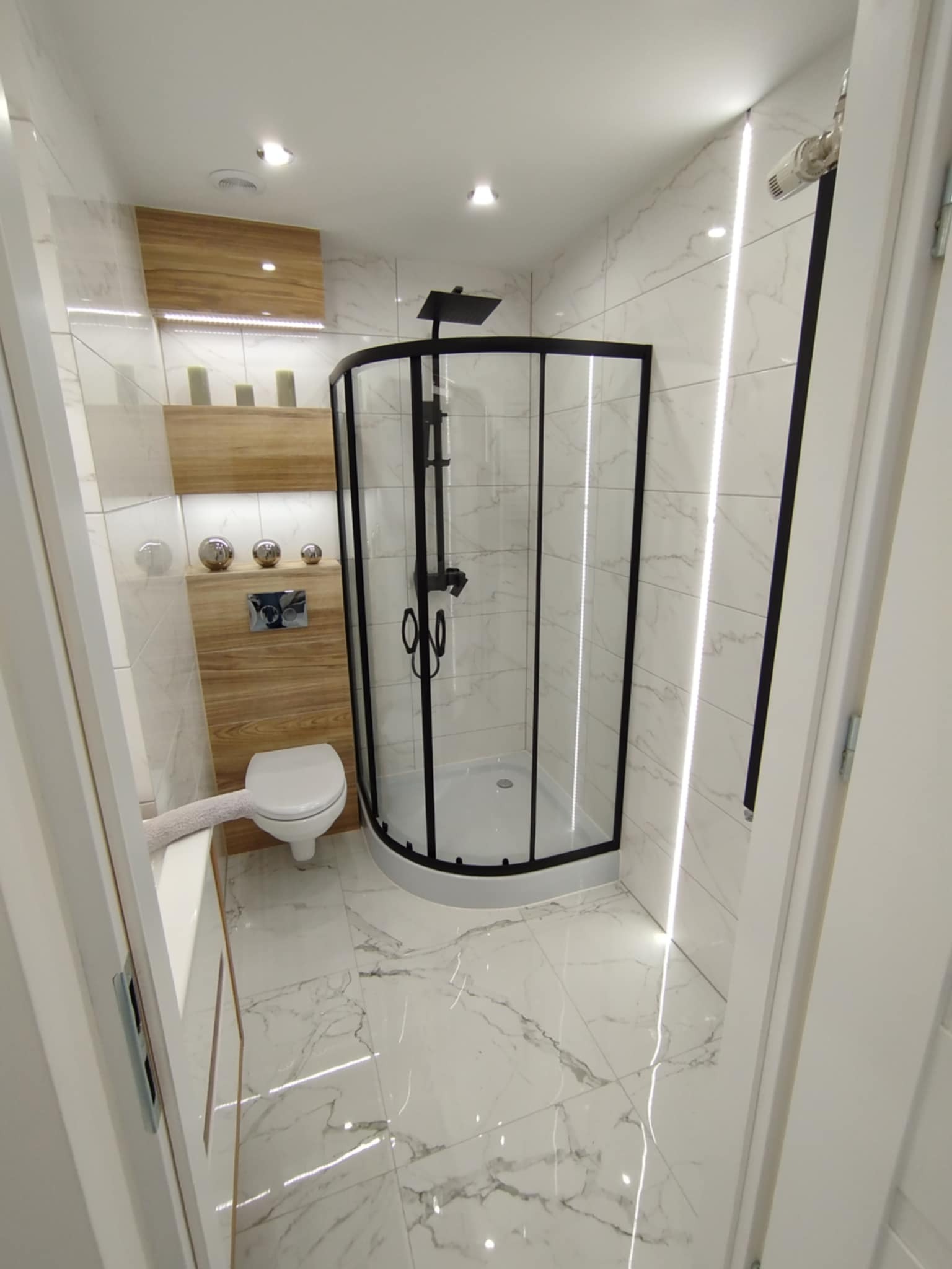 Wykończona łazienka z kabiną prysznicową w czarnej ramie, toaletą podwieszaną i marmurowymi płytkami, oświetlona ledami. Drewniane elementy dekoracyjne.