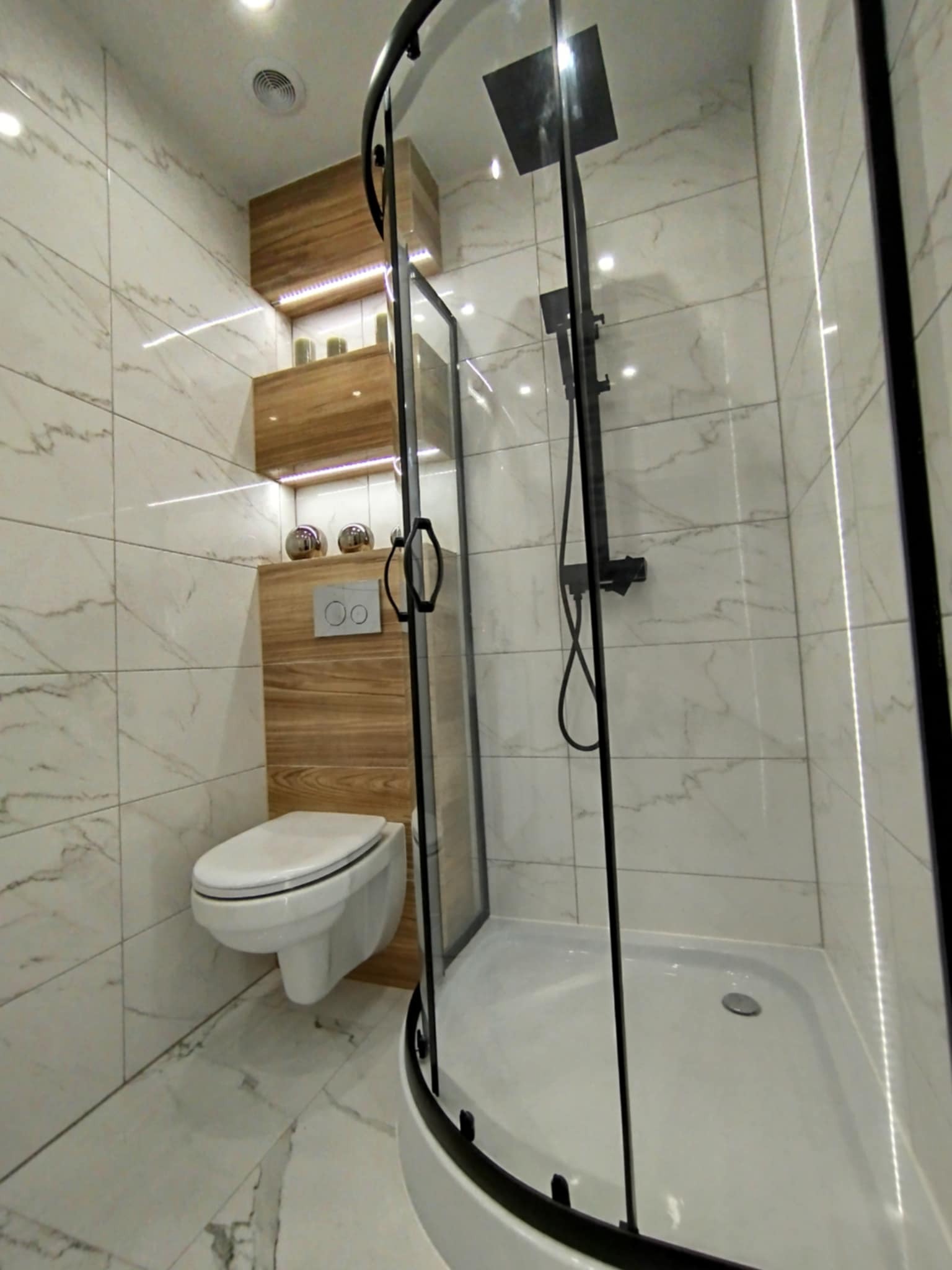 Nowoczesna łazienka z kabiną prysznicową typu walk-in, podwieszaną toaletą i drewnianymi półkami z oświetleniem LED, wykończona marmurowymi płytkami.
