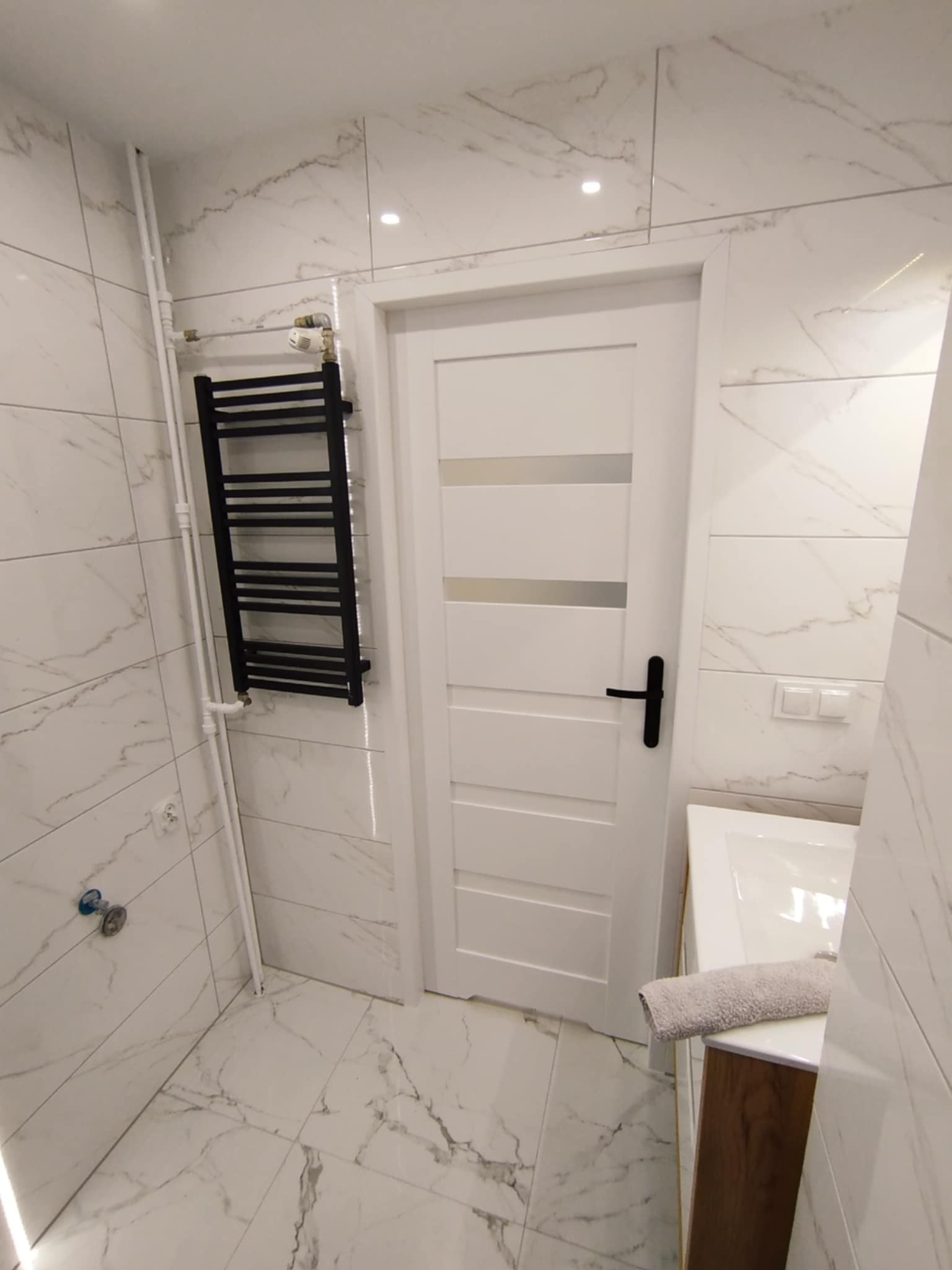 Elegancka łazienka z białymi marmurkowymi płytkami, czarnym grzejnikiem drabinkowym i białą stolarką drzwiową z czarną klamką. Minimalistyczny design.
