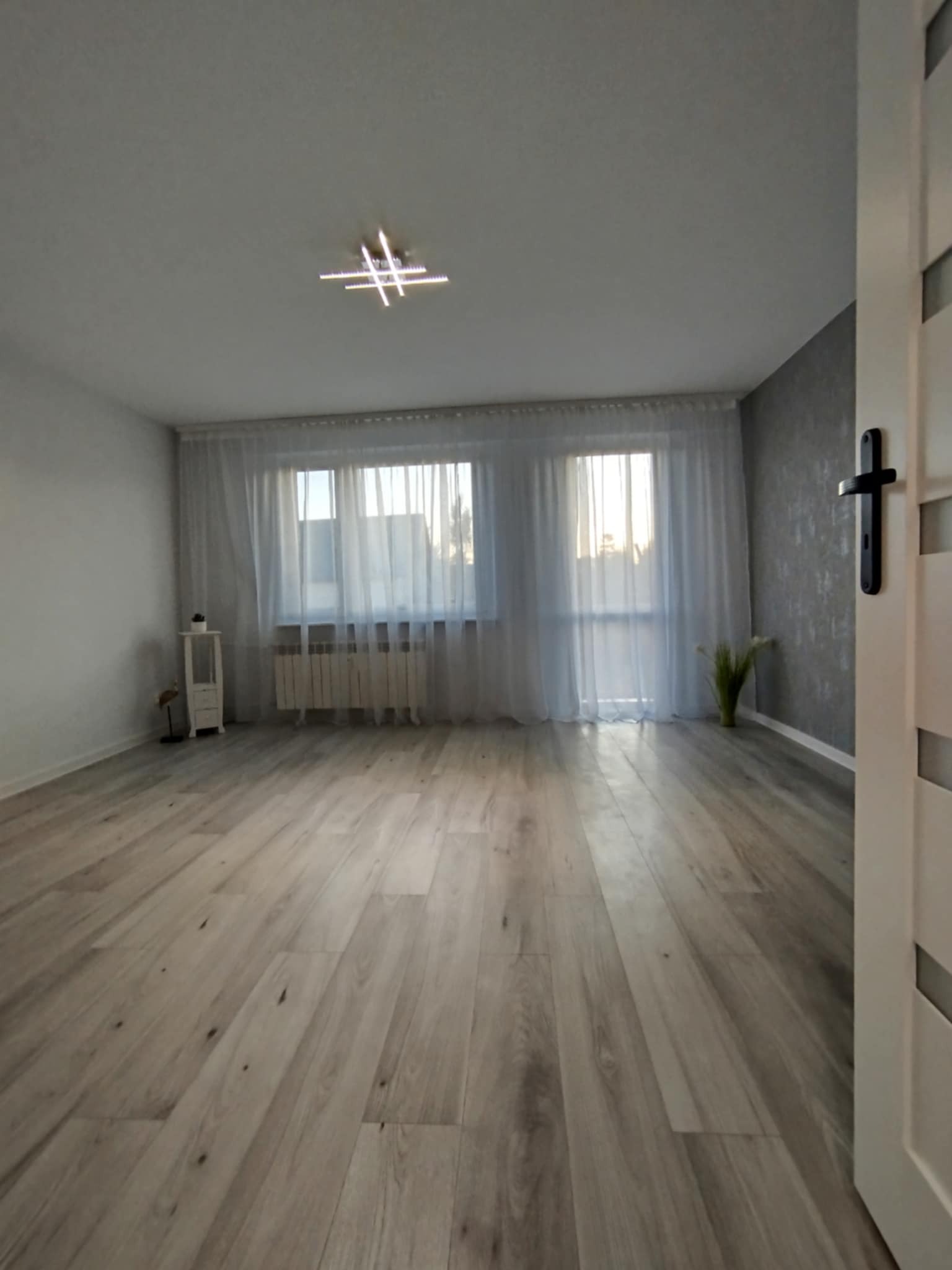 Jasne, przestronne wnętrze po remoncie: panele podłogowe imitujące drewno, białe ściany, okno z firankami i nowoczesna lampa sufitowa.