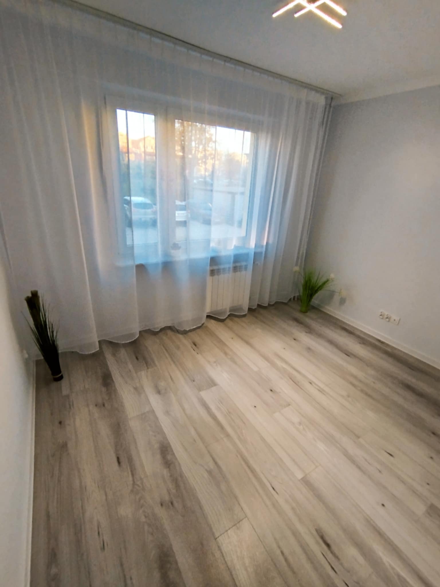Jasne panele podłogowe w pokoju z oknem przysłoniętym białymi firankami. Widoczny grzejnik pod oknem oraz dekoracyjne trawy w doniczkach. Minimalistyczne oświetlenie sufitowe.