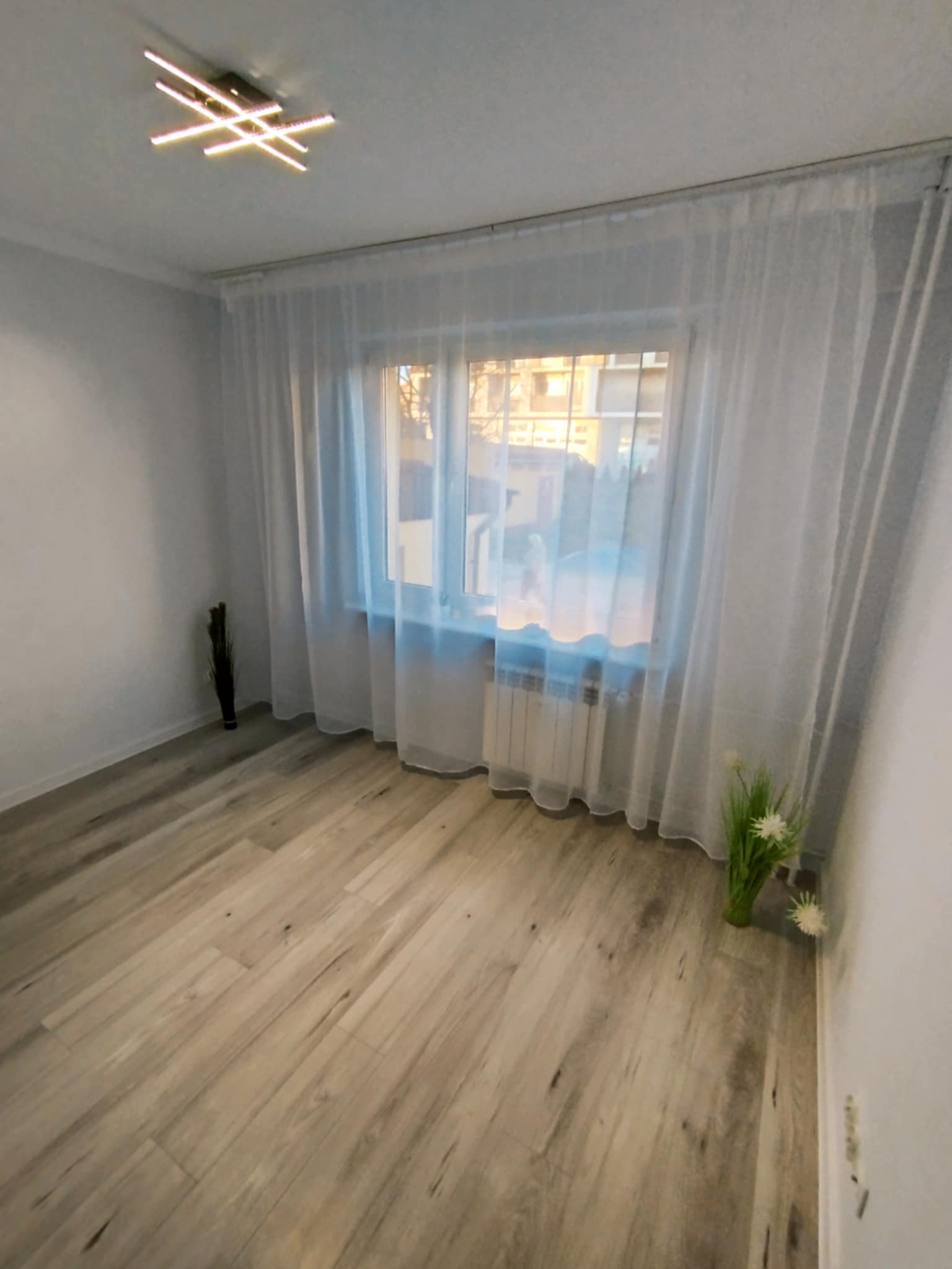 Jasne panele podłogowe w pokoju z oknem przysłoniętym białymi firankami. Na suficie nowoczesna, geometryczna lampa. Minimalistyczny wystrój wnętrza.