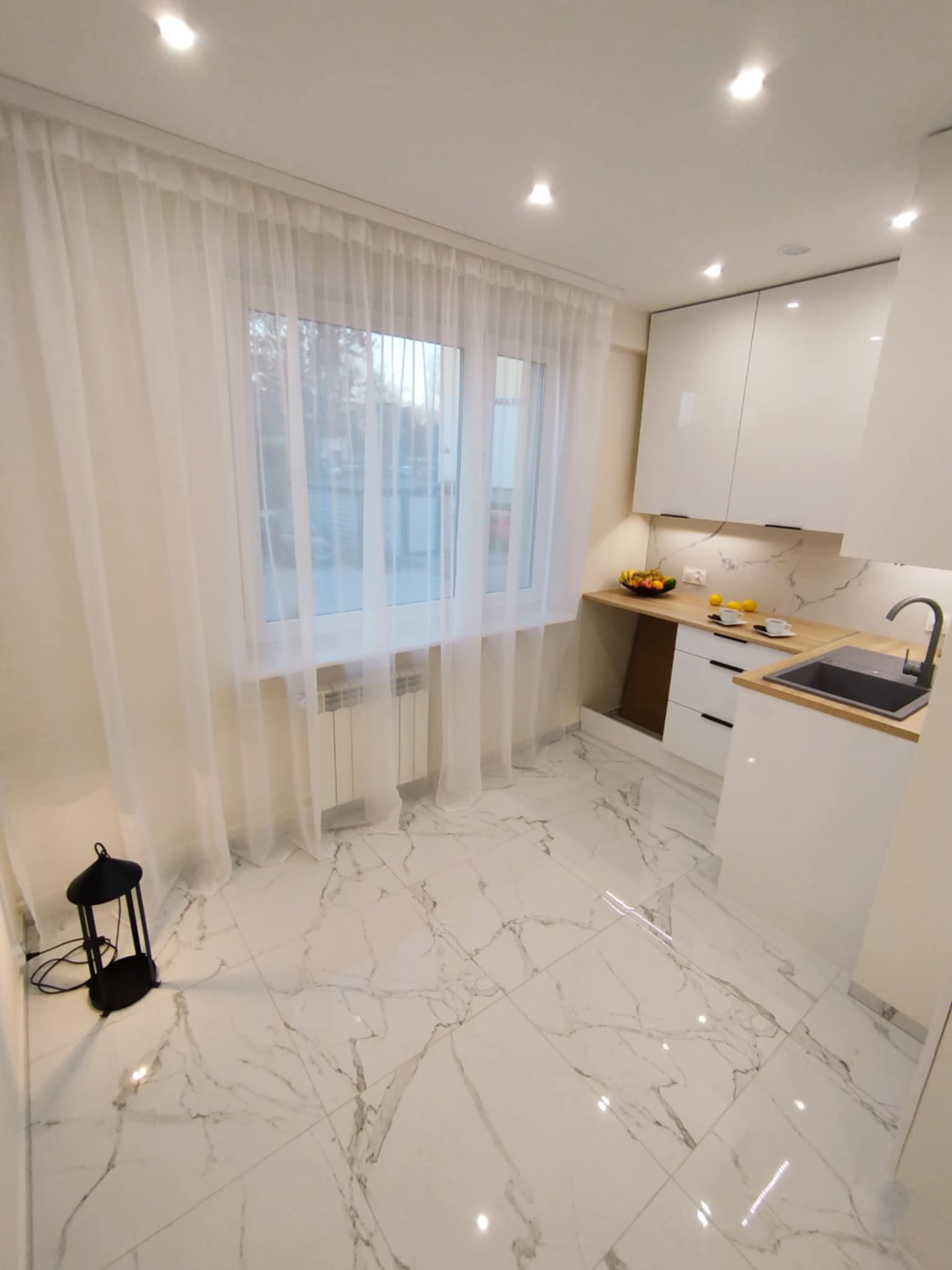 Biała kuchnia z marmurową podłogą i blatem, minimalistyczny design. Widok na okno z firanką, czarna lampa w rogu. Nowoczesne oświetlenie.